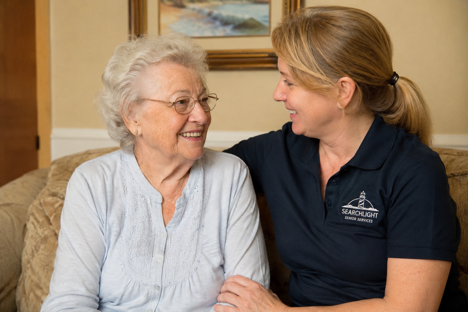 An elderly woman and a caregiver smiling at each other indoors, sitting on a sofa.