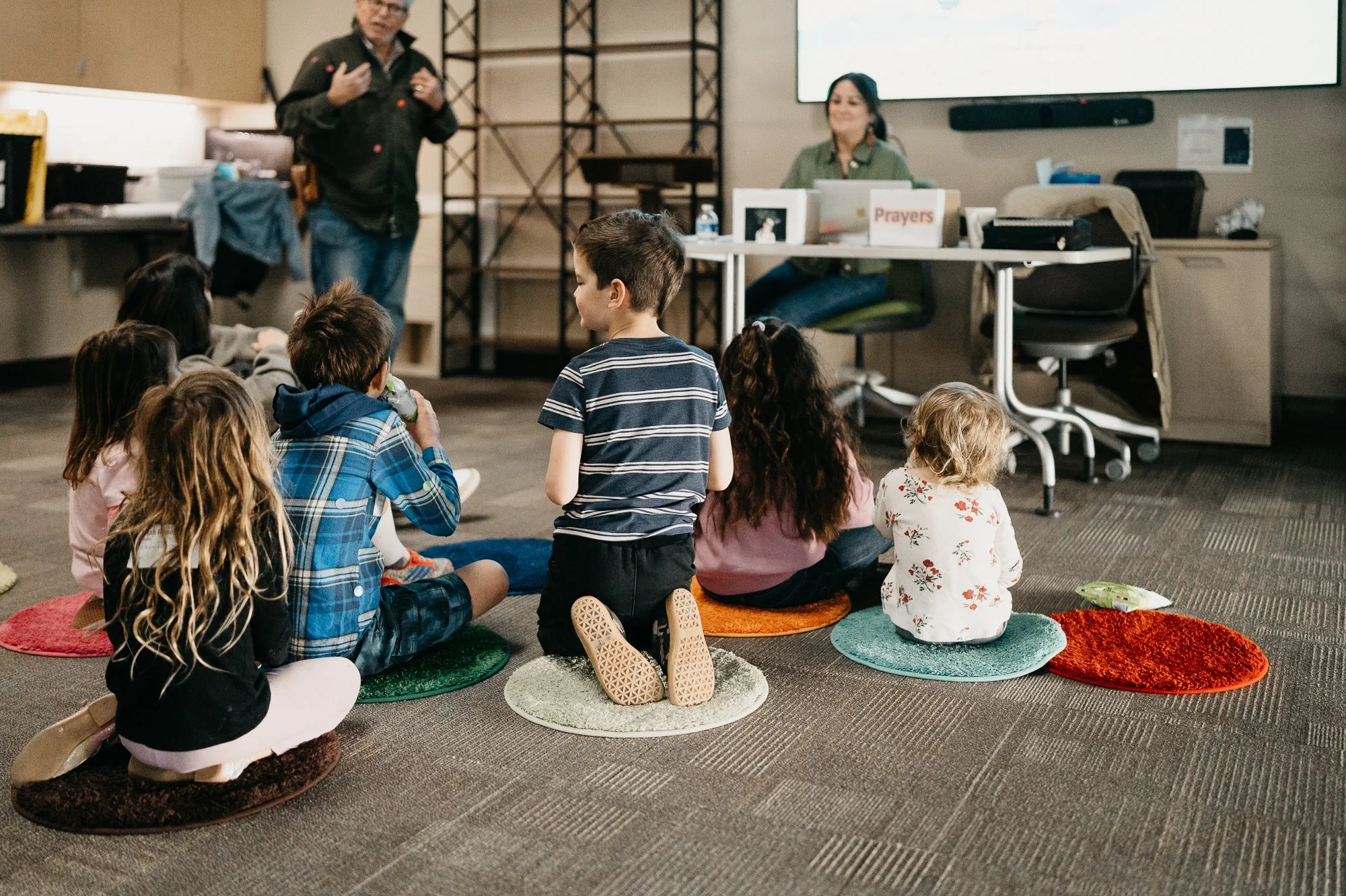 Children sitting on colorful cushions listening to a speaker in a classroom or community center setting.