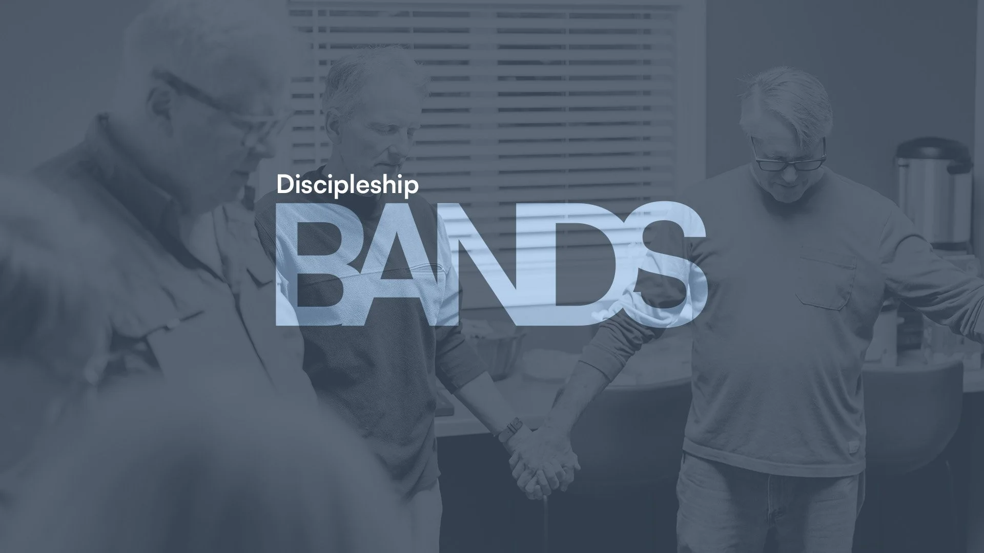 A group of four men holding hands in a prayer circle in a room with blinds.