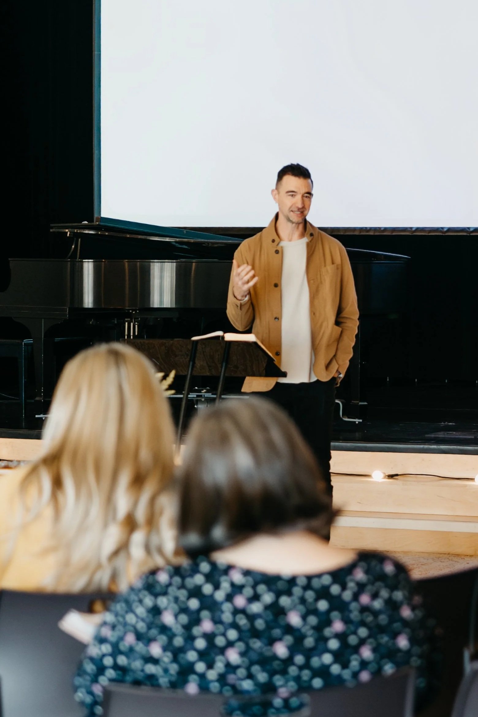 Jason Mayer giving a presentation or speech in front of an audience, standing by a grand piano, with people listening.