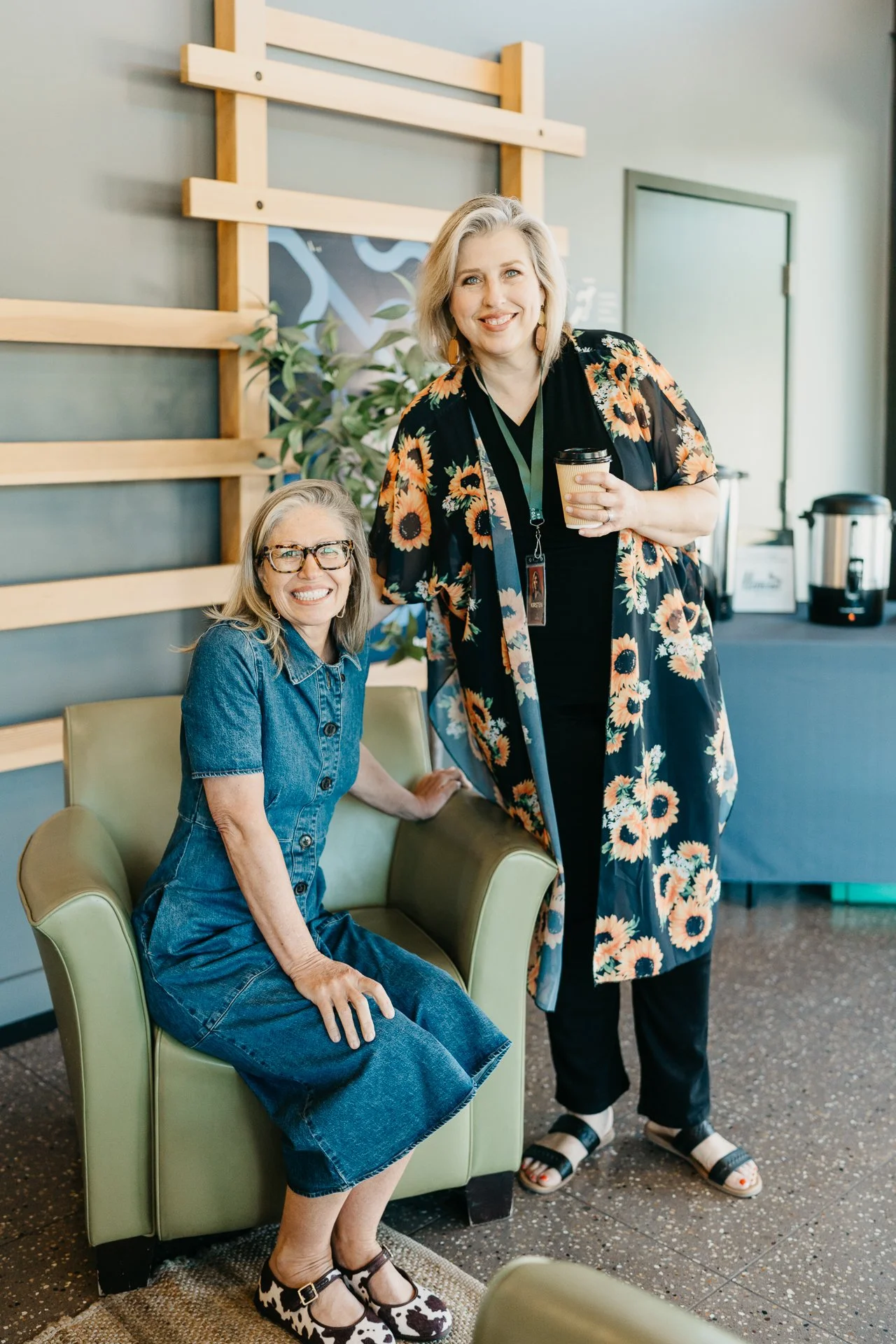 Two women smiling in a casual indoor setting, one sitting on a green armchair and the other standing with a coffee cup, with a decorative wall and greenery in the background.