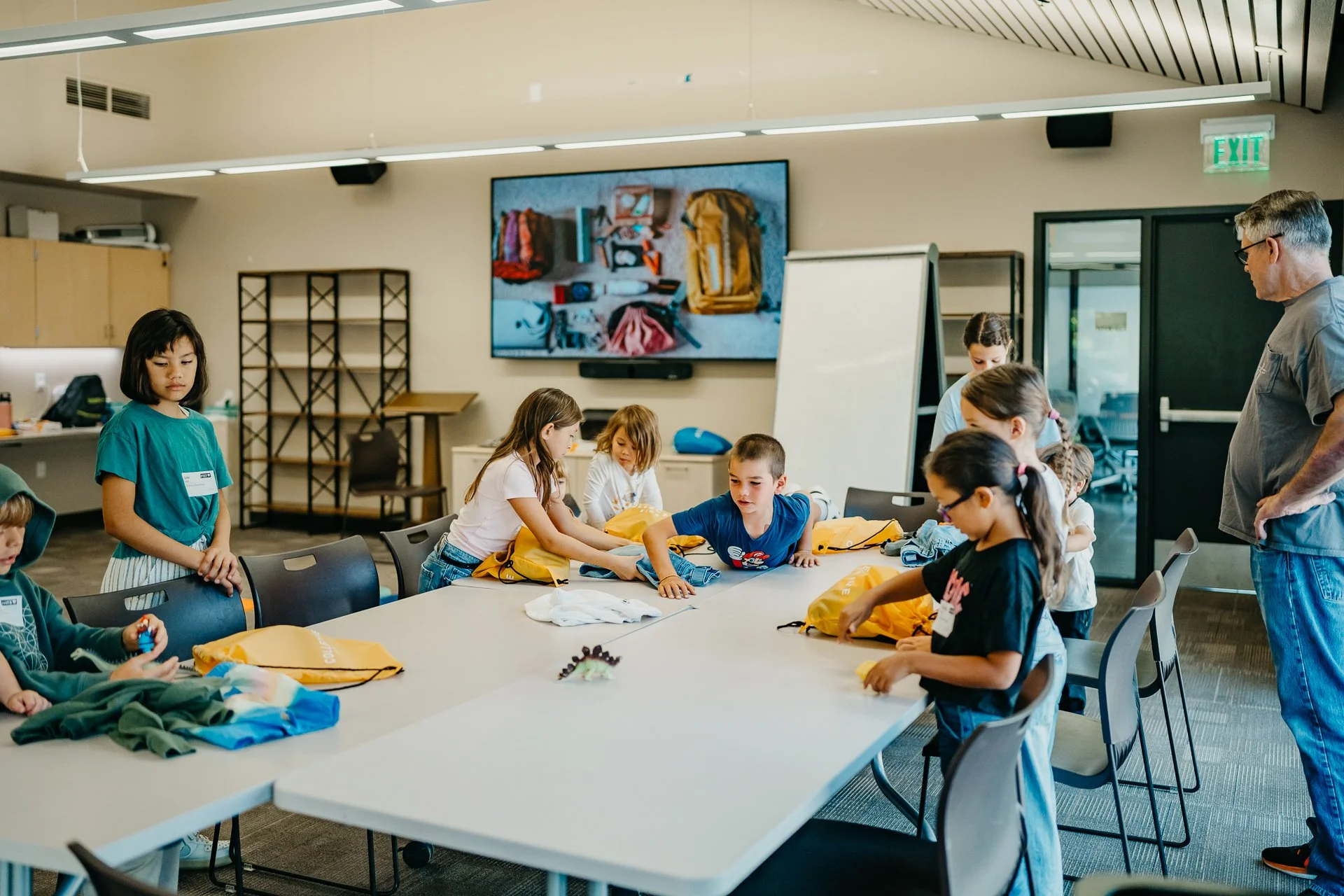 Collective Kids Children and adults in a classroom or workshop setting around a large table with yellow backpacks, preparing for an activity.