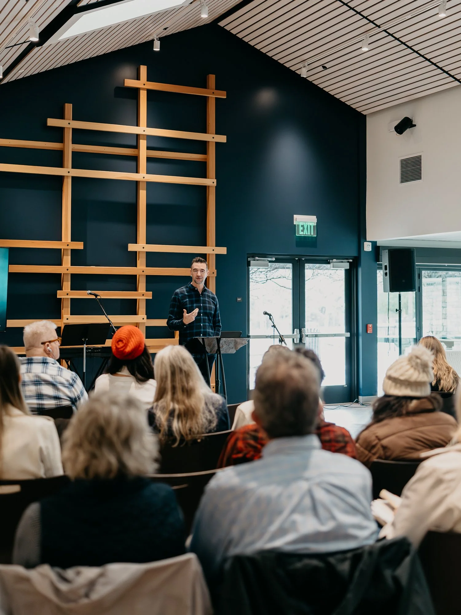 A man giving a presentation on stage in front of an audience in a church or auditorium with a large wall featuring wooden cross-shaped decor.