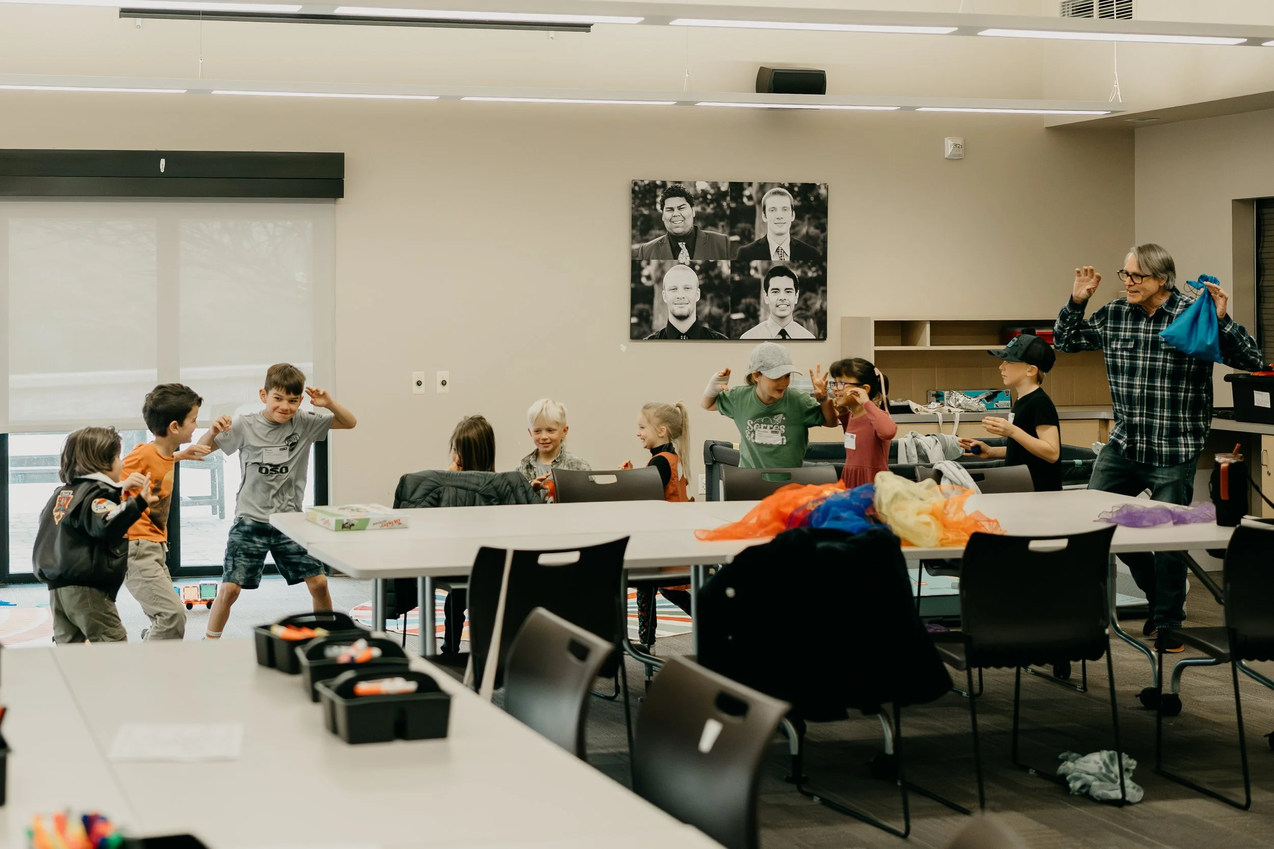 A group of children and an adult in a brightly lit classroom, engaging in playful activities around tables with colorful supplies, with a large window on the left and black and white portraits on the wall.