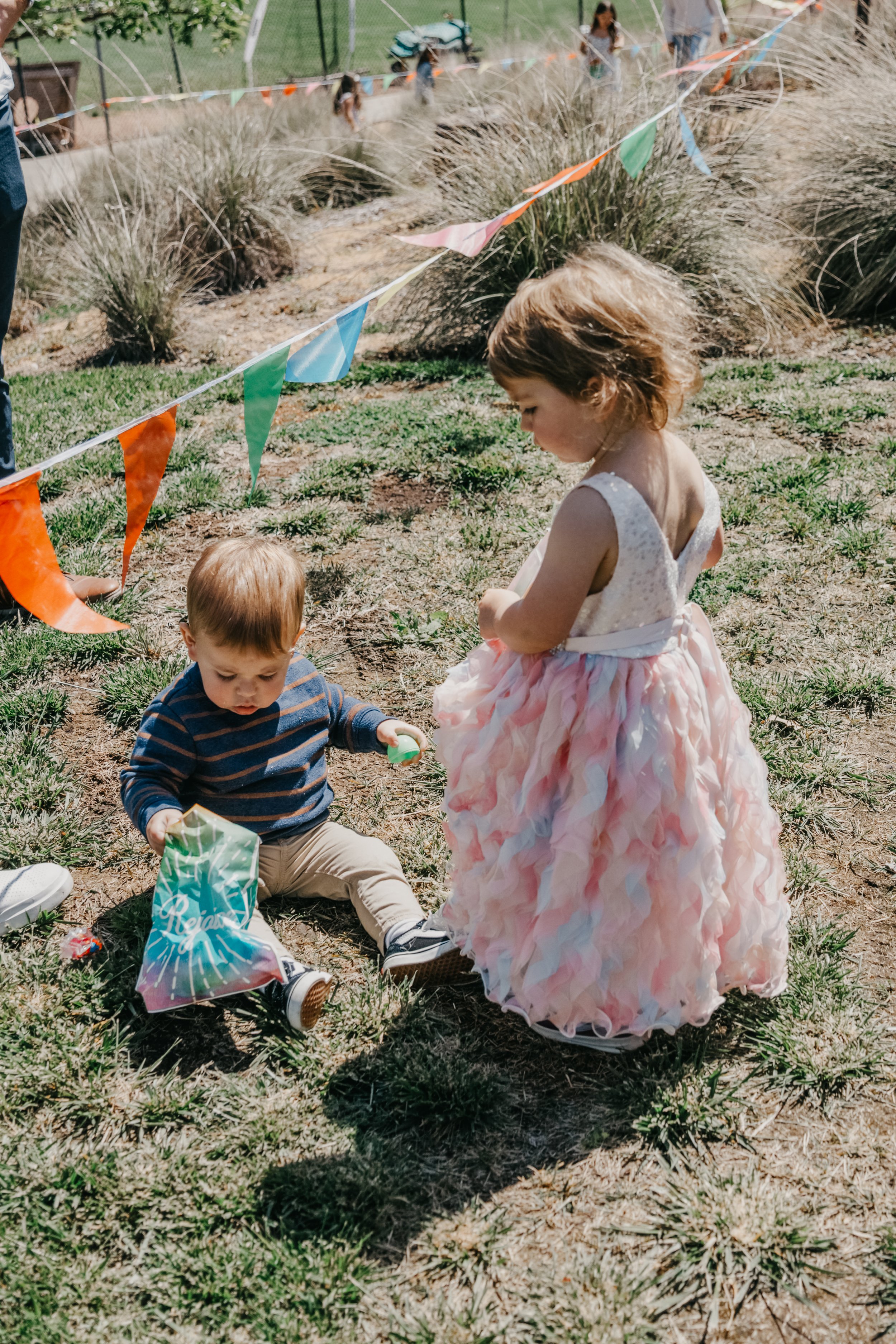 Two young children, a boy and a girl, playing outdoors at a festive event decorated with colorful bunting. The boy is sitting on the ground, holding a party hat and a small toy, while the girl stands nearby in a pink and white dress, looking down at him.