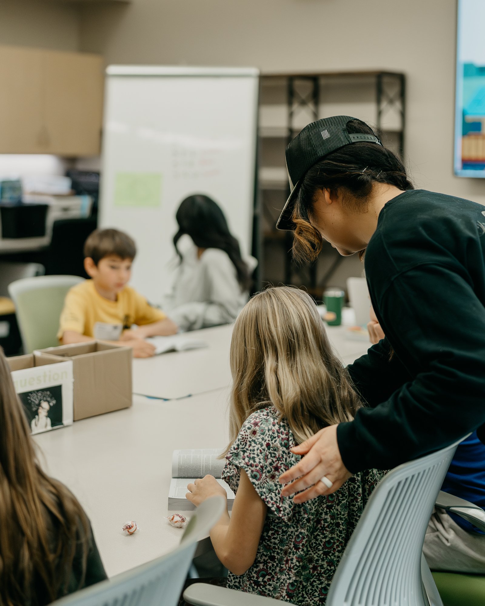 A woman in a black shirt and cap leaning over a girl with long blonde hair reading a book at a table in a classroom or learning environment.