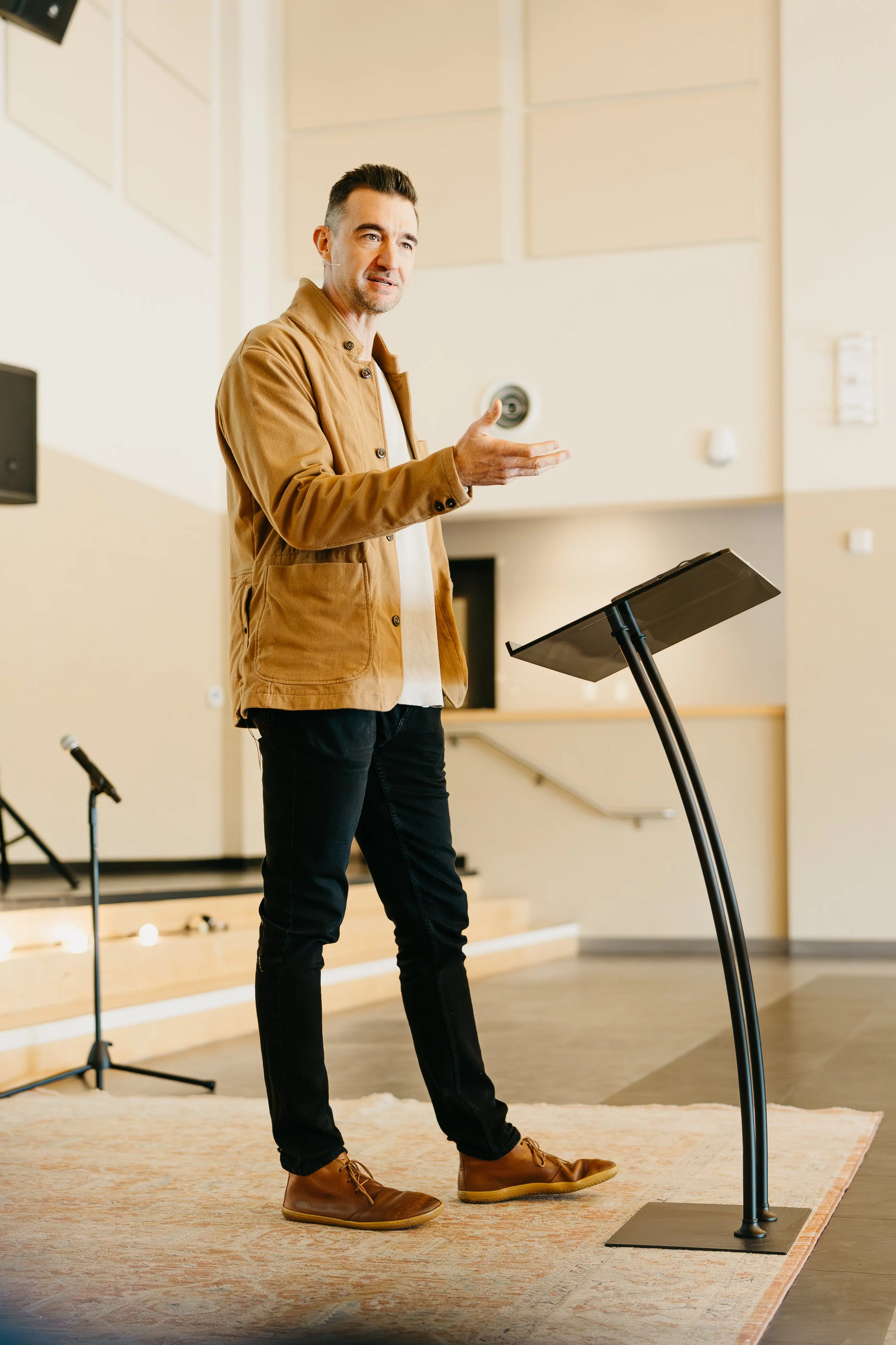 Man giving a presentation in a lecture hall or conference room, standing near a lectern, gesturing with his hand.