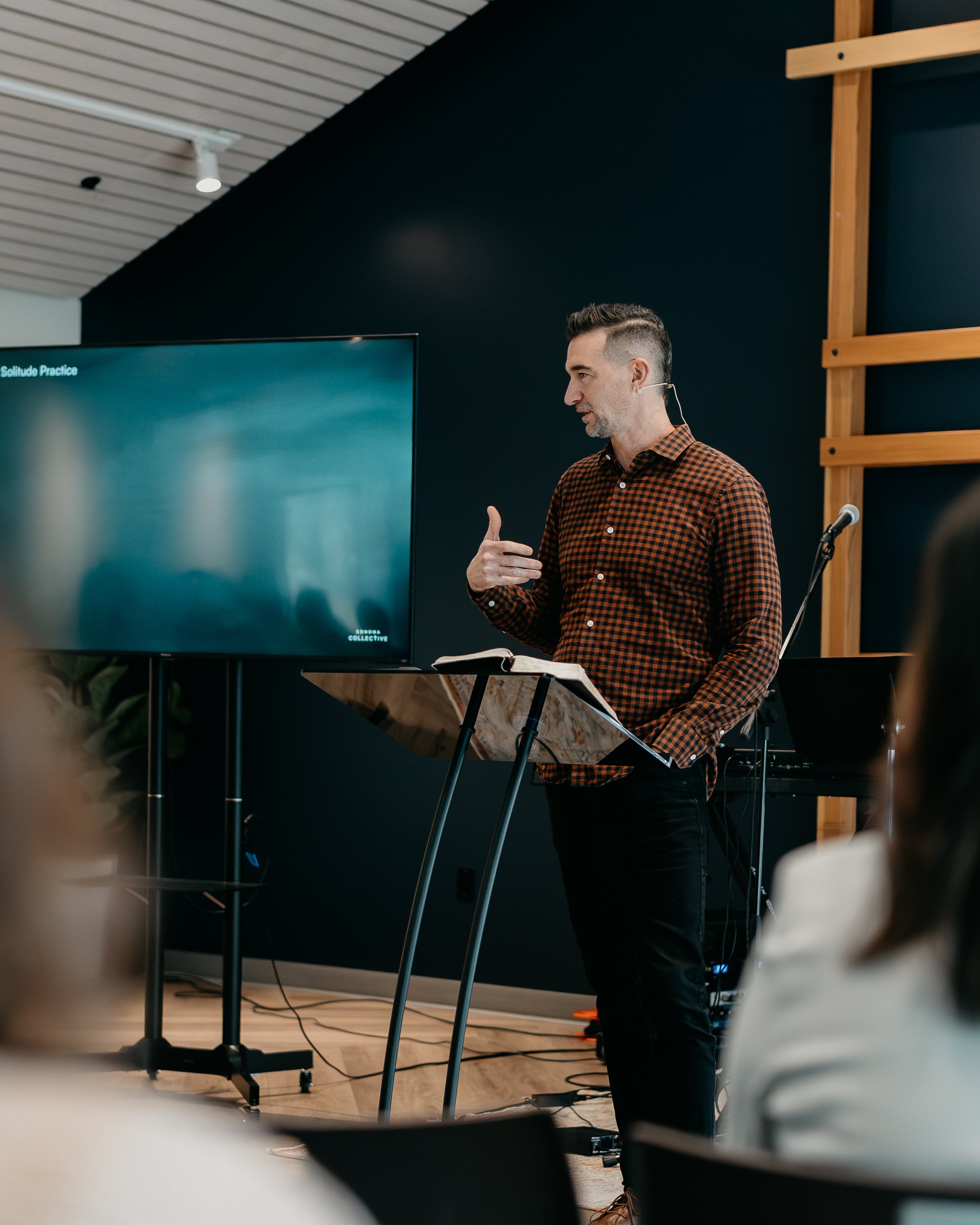 A man giving a presentation in front of a large screen at a conference or classroom setting, with an open book on a stand, people in the audience are partially visible.