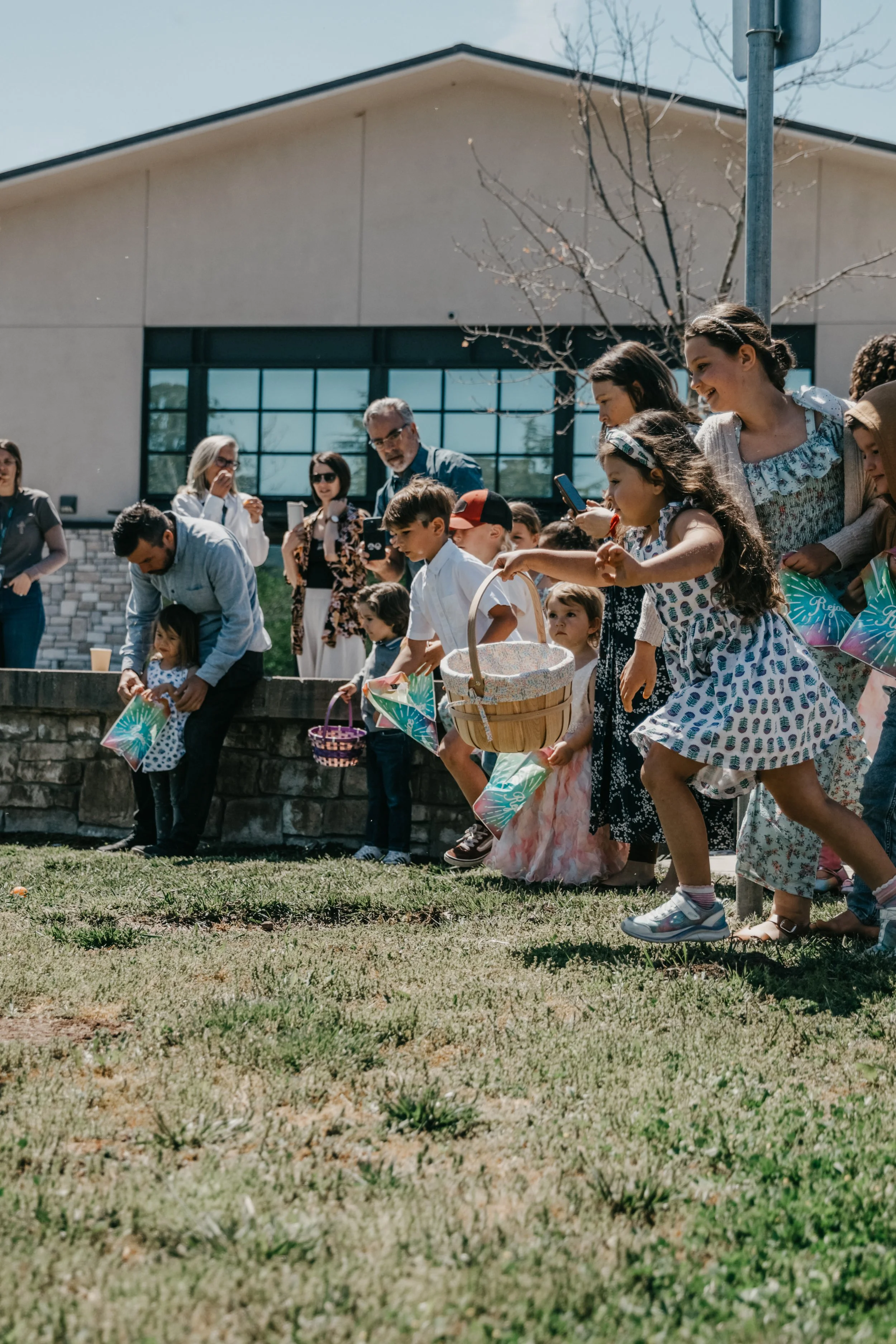 Children and adults participate in an outdoor egg hunt, with some children holding baskets, at a sunny day on a grassy area near a building with large windows.