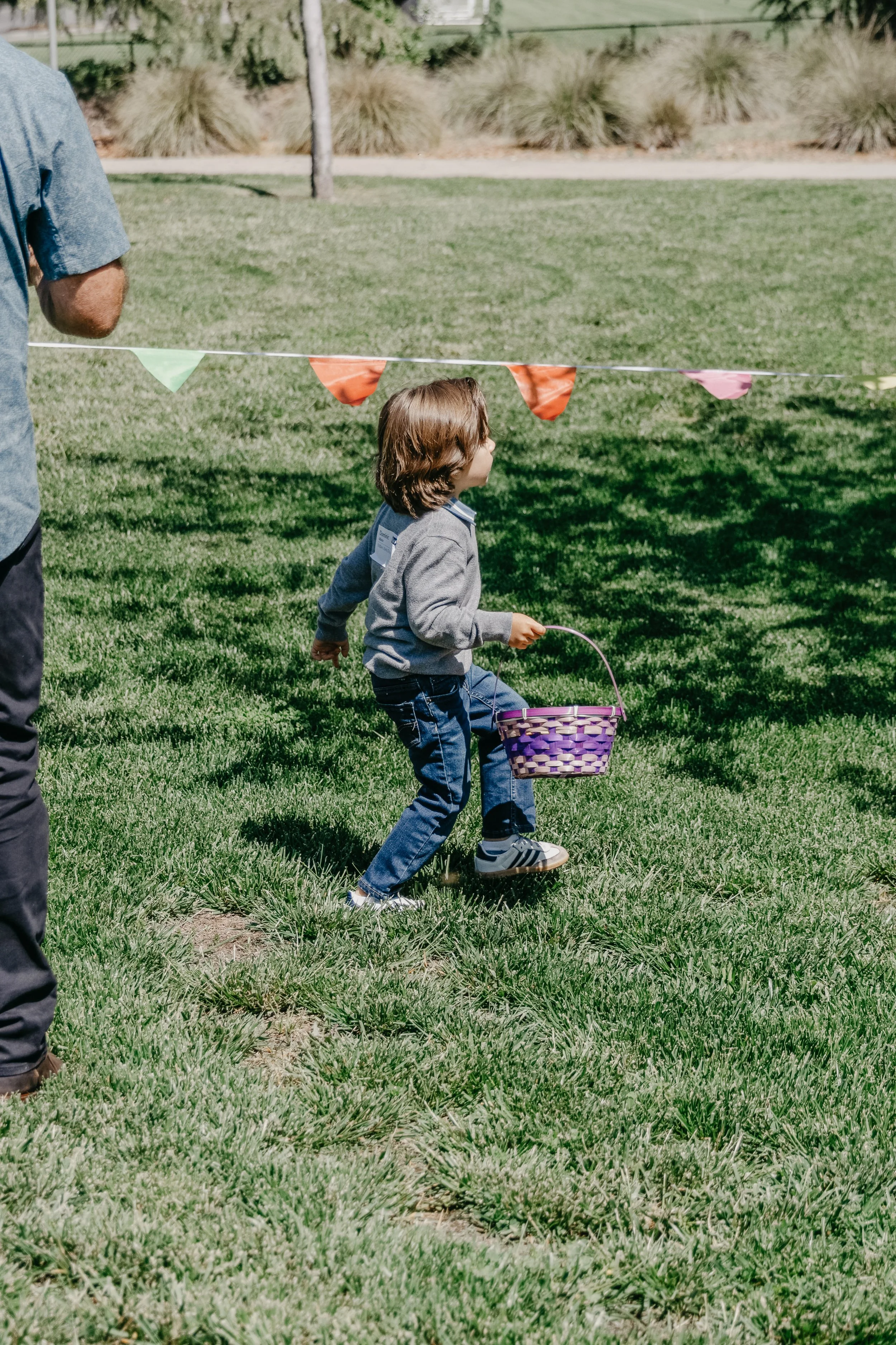 A young boy holding a purple basket, walking under colorful pennant banners in a grassy park.