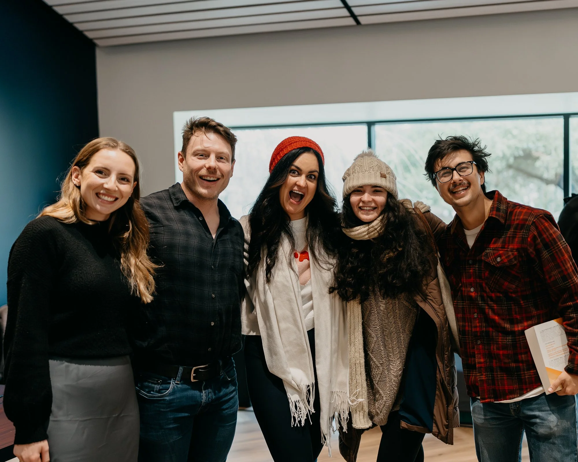 Group of five smiling young adults embracing each other indoors near a large window.