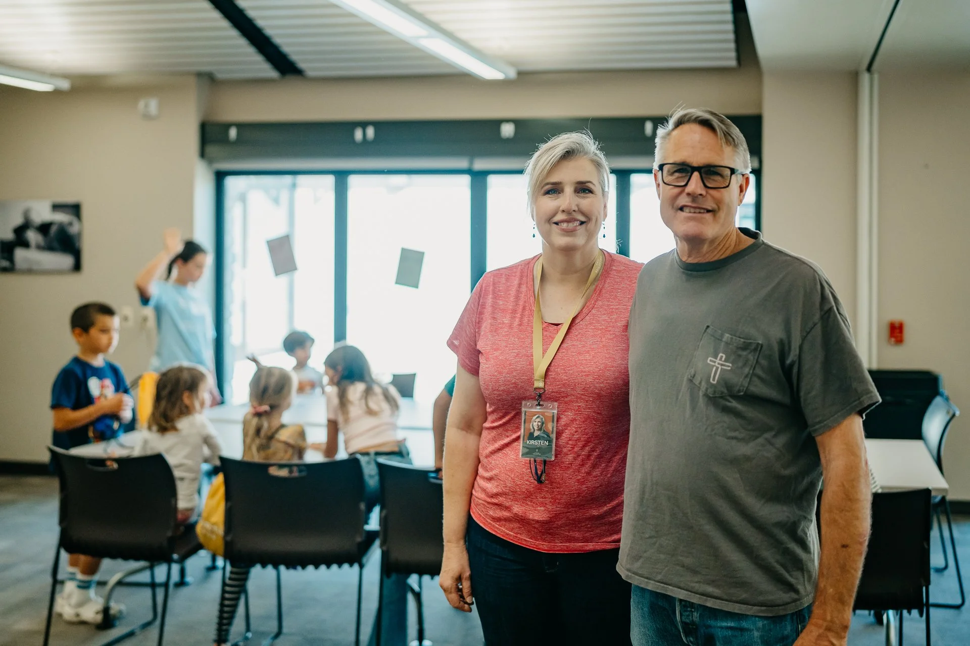 Two adults smiling inside a classroom, with children sitting at tables in the background.