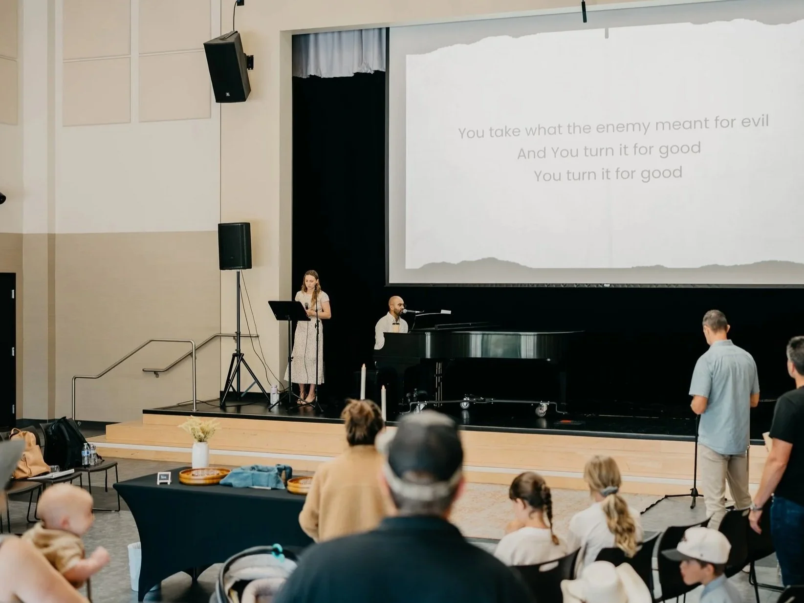 A church or conference hall with a stage featuring a woman singing with a microphone, a man playing a grand piano, and a large screen displaying song lyrics. Audience members are seated and standing, watching the performance.