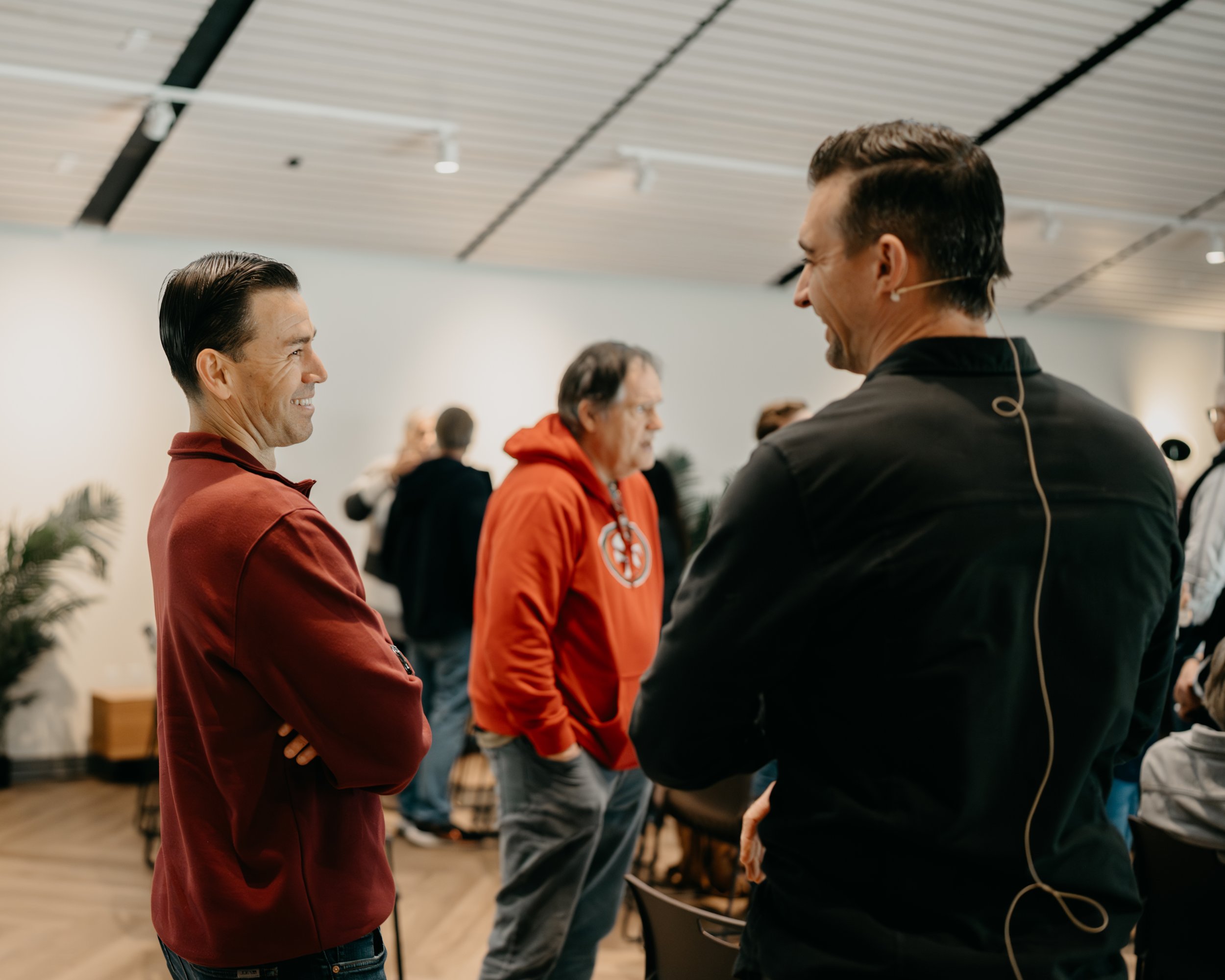 Two men are talking and smiling in a room with several people in the background. One man is wearing a red jacket, and the other is dressed in black. The room has a modern ceiling and some plants.