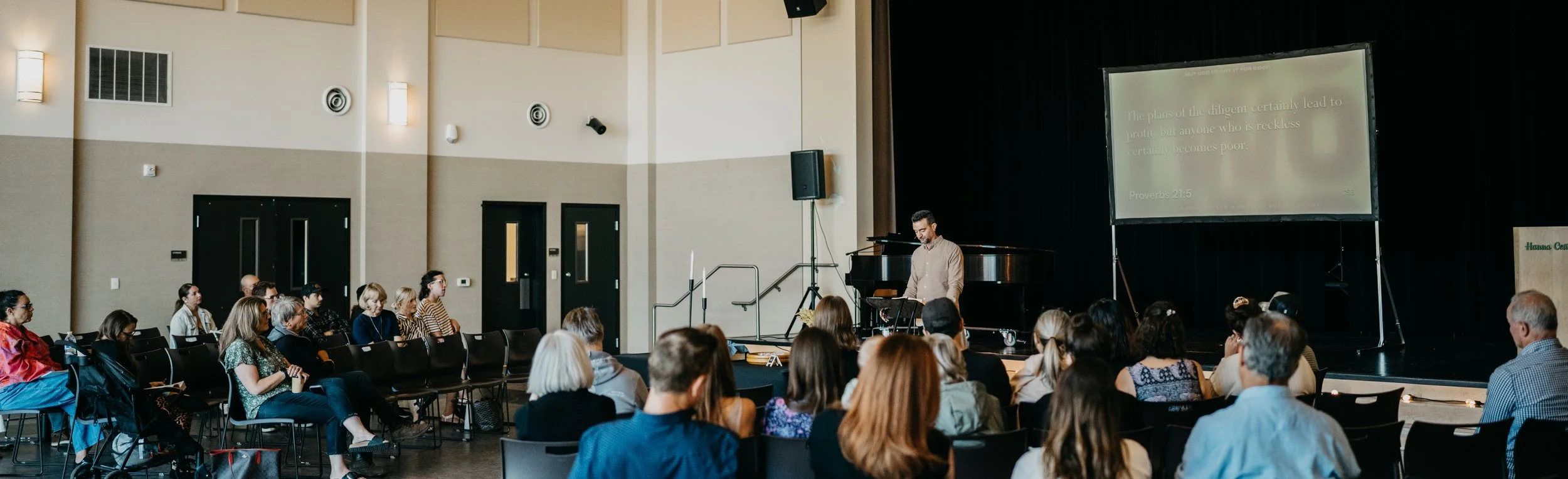 A man presenting on stage in an auditorium with an audience seated and watching. The stage has a large screen displaying a quote from Proverbs 21:5.
