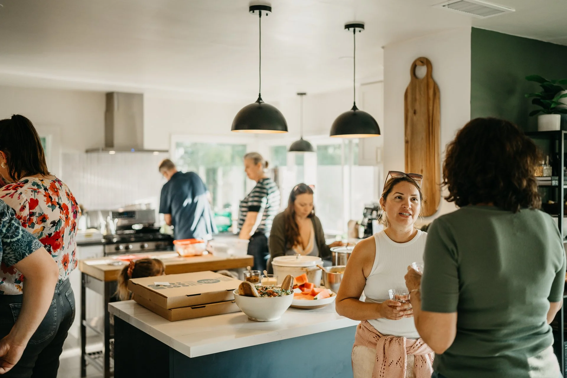 People gathered in a modern kitchen, engaging in conversation and preparing food on a white island with pizza boxes, bowls of food, and other kitchen items, with black pendant lights hanging from the ceiling.