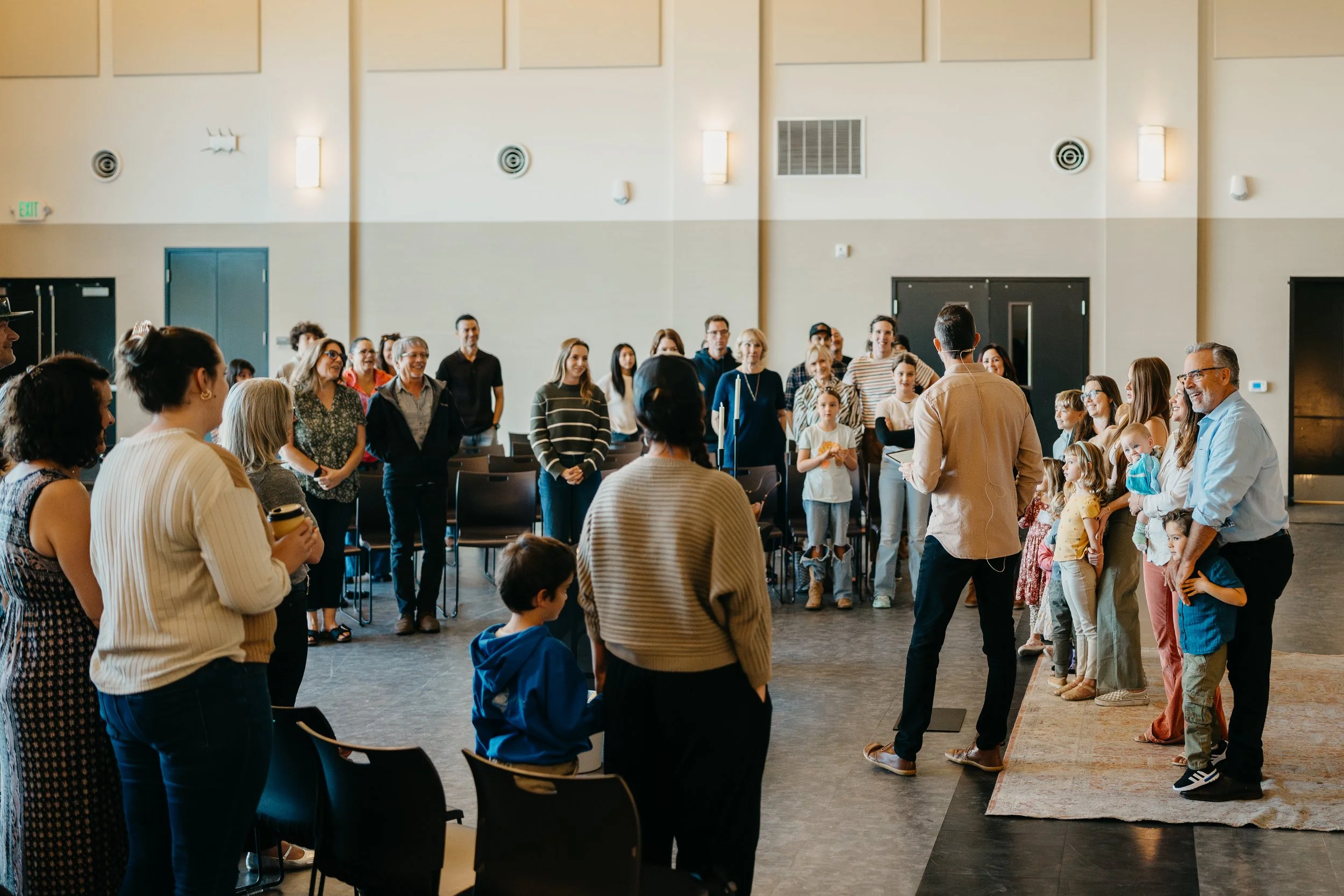 A group of people (Sonoma Collective) gathered in a large, well-lit room, standing and sitting, listening to a speaker standing on a rug at the front. The group includes children, adults, and seniors, with some smiling and engaged.