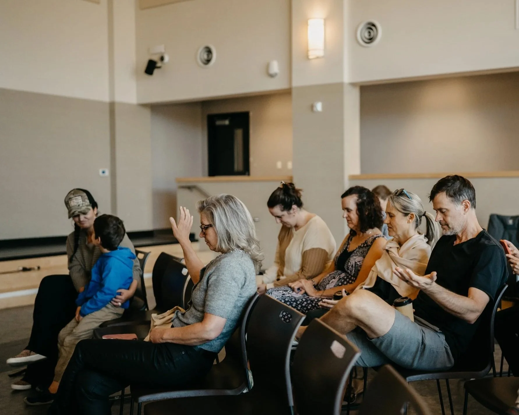 Group of people sitting on chairs in a room, some with eyes closed, appearing to meditate or pray.