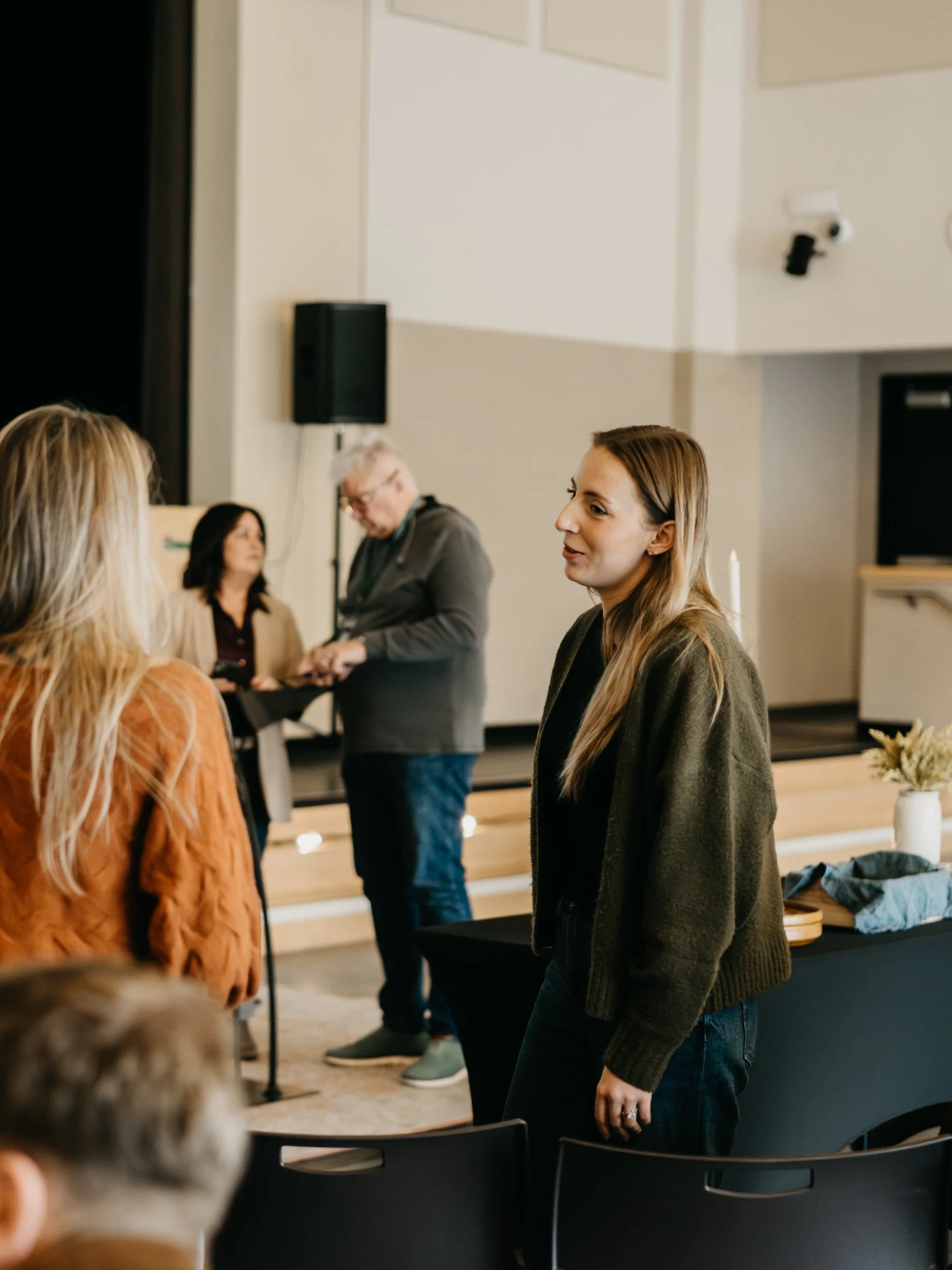 A woman with long, light brown hair stands and talks in a conference room with several people. In the background, a man and a woman are near a speaker and some presentation equipment.