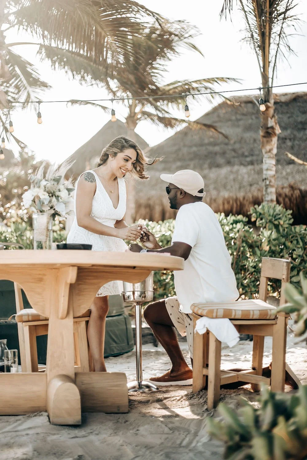 A woman in a white dress holds hands with a man in a white shirt and cap, sitting at a beachside wooden table in Tulum. Palm trees and fairy lights adorn the background, creating a romantic setting.