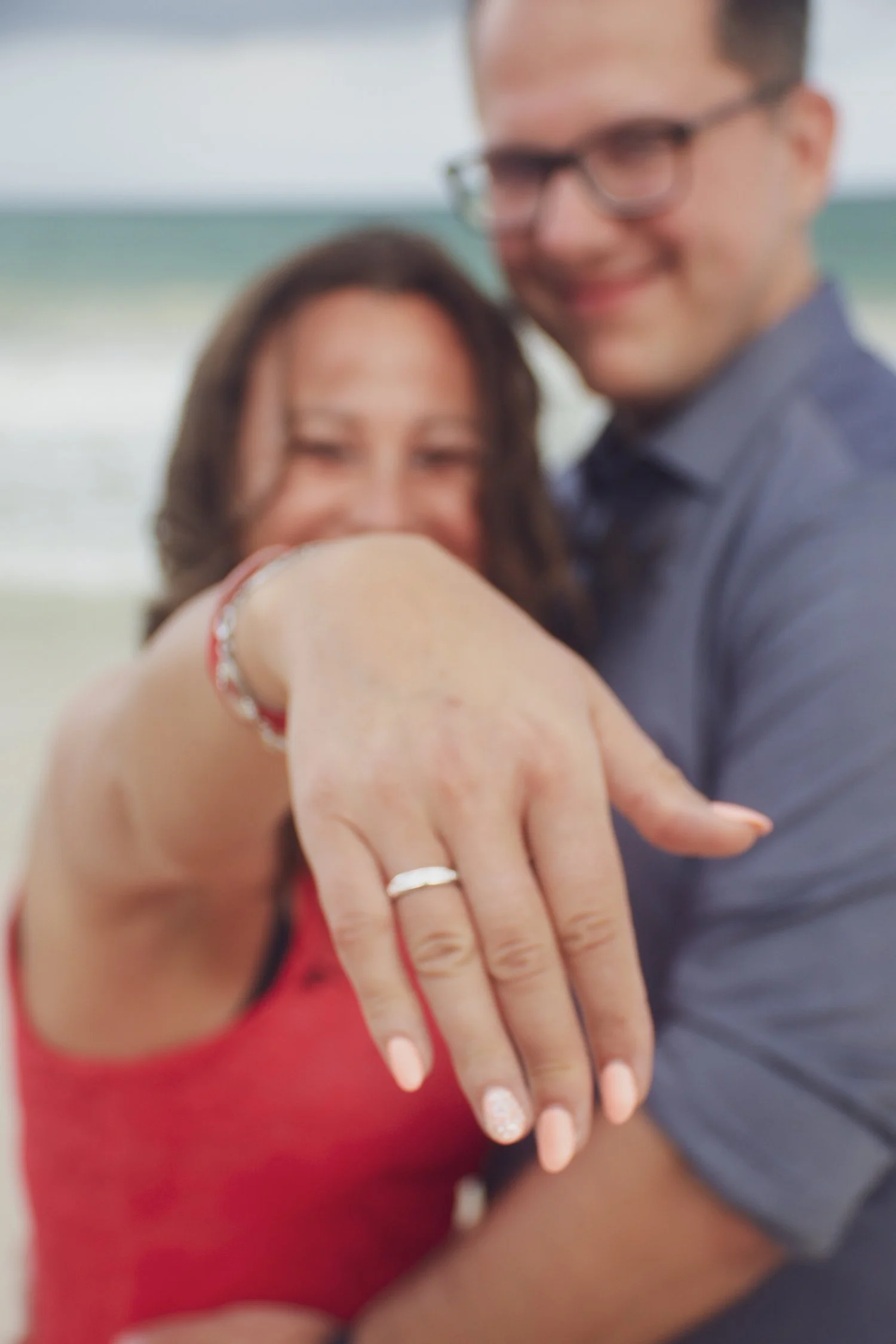 A couple smiling, with the woman prominently displaying an engagement ring on her hand, set against a beach background. Custom proposal experience Mexico.