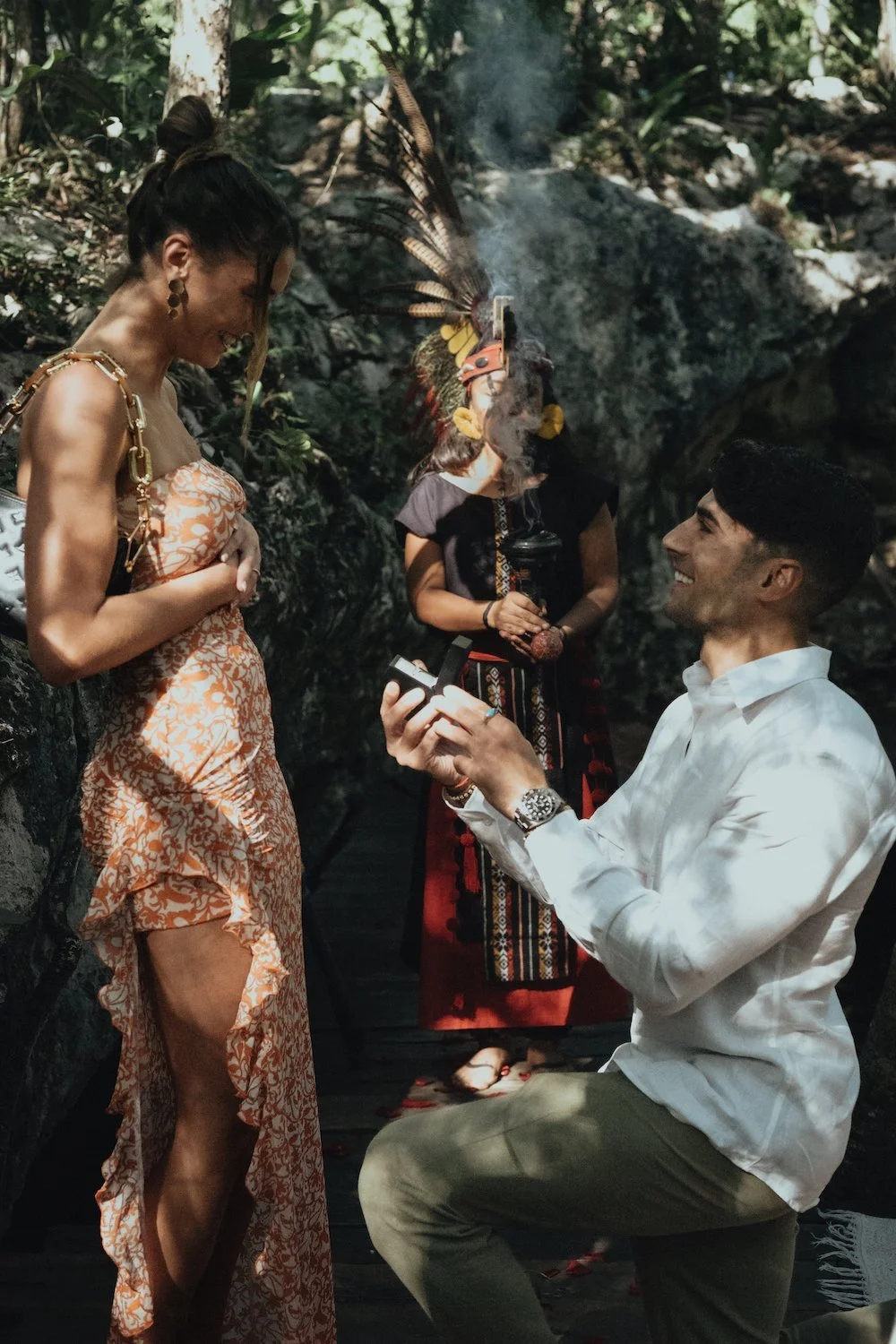 Man proposing to woman in a forest setting with a ceremonial figure holding incense nearby planned by a luxury marriage proposal agency in Tulum.