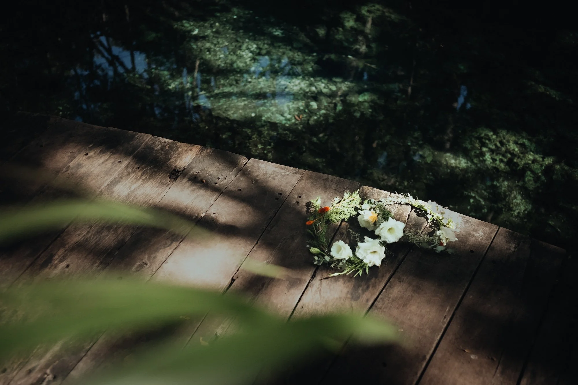 A floral wreath with white and orange flowers lies on a wooden deck near a pond with reflections of surrounding trees.