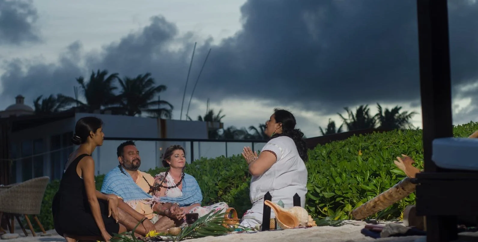 A group of four people sitting in a circle outside, engaging in a marriage proposal ceremony. They are on a sandy surface, surrounded by greenery, with dark clouds and palm trees in the background.
