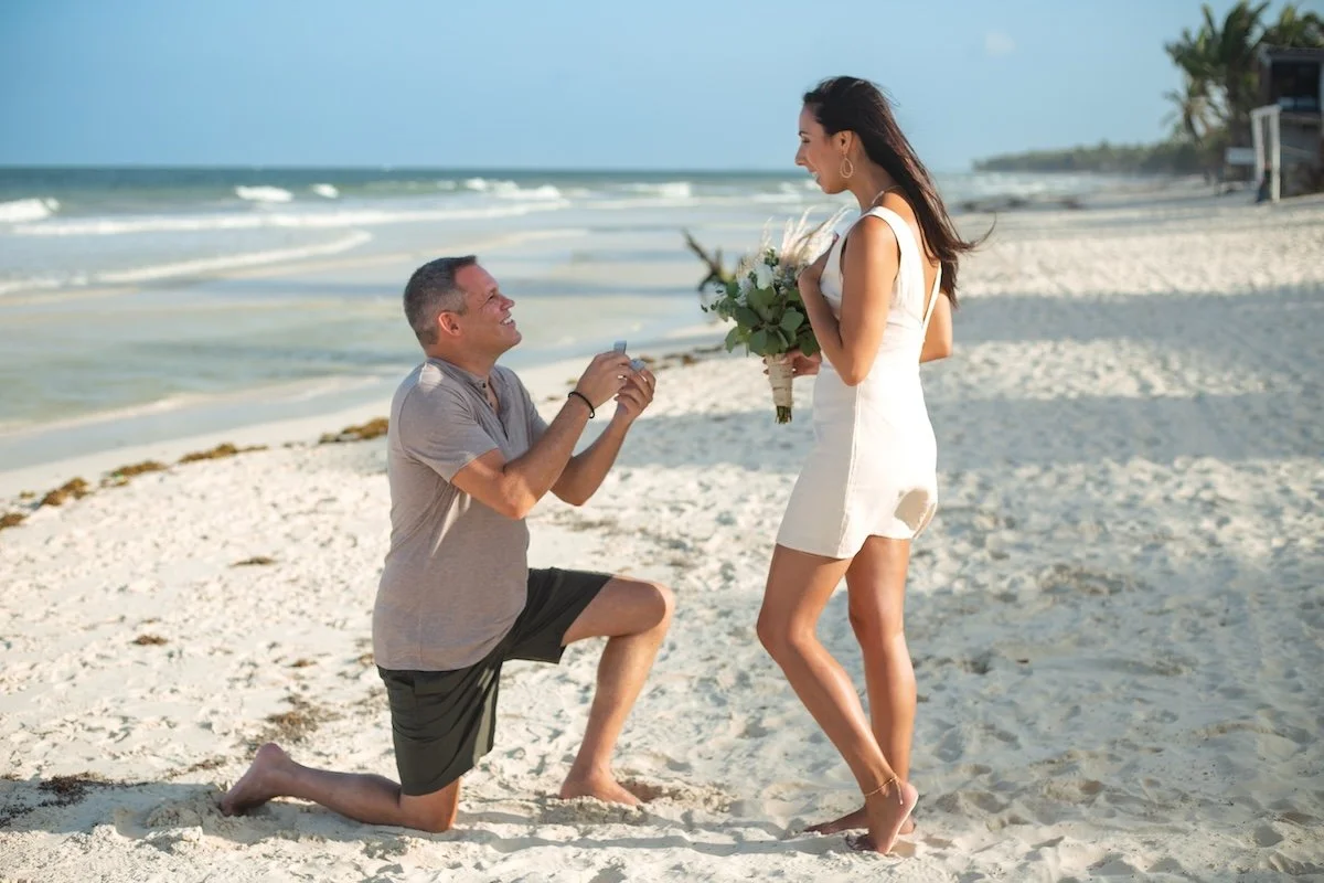 Man proposing to woman on beach, holding a ring box, woman with bouquet, romantic proposal Tulum. 