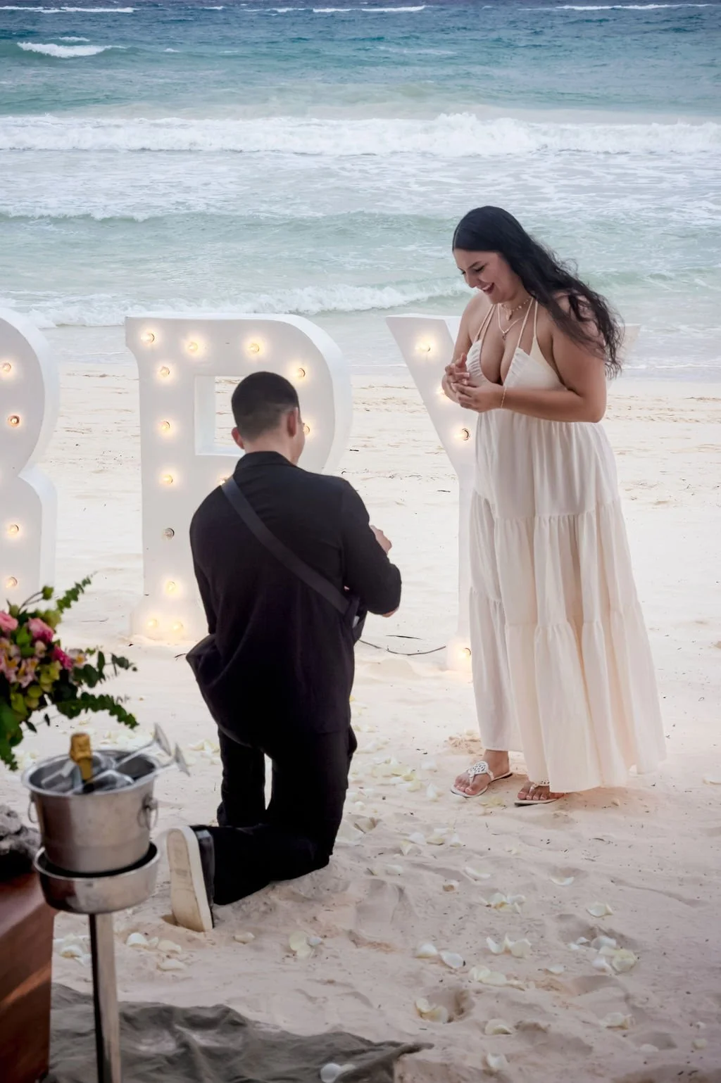 Man proposing to woman on a beach in Tulum, surrounded by large lit-up letters and flower petals on the sand.