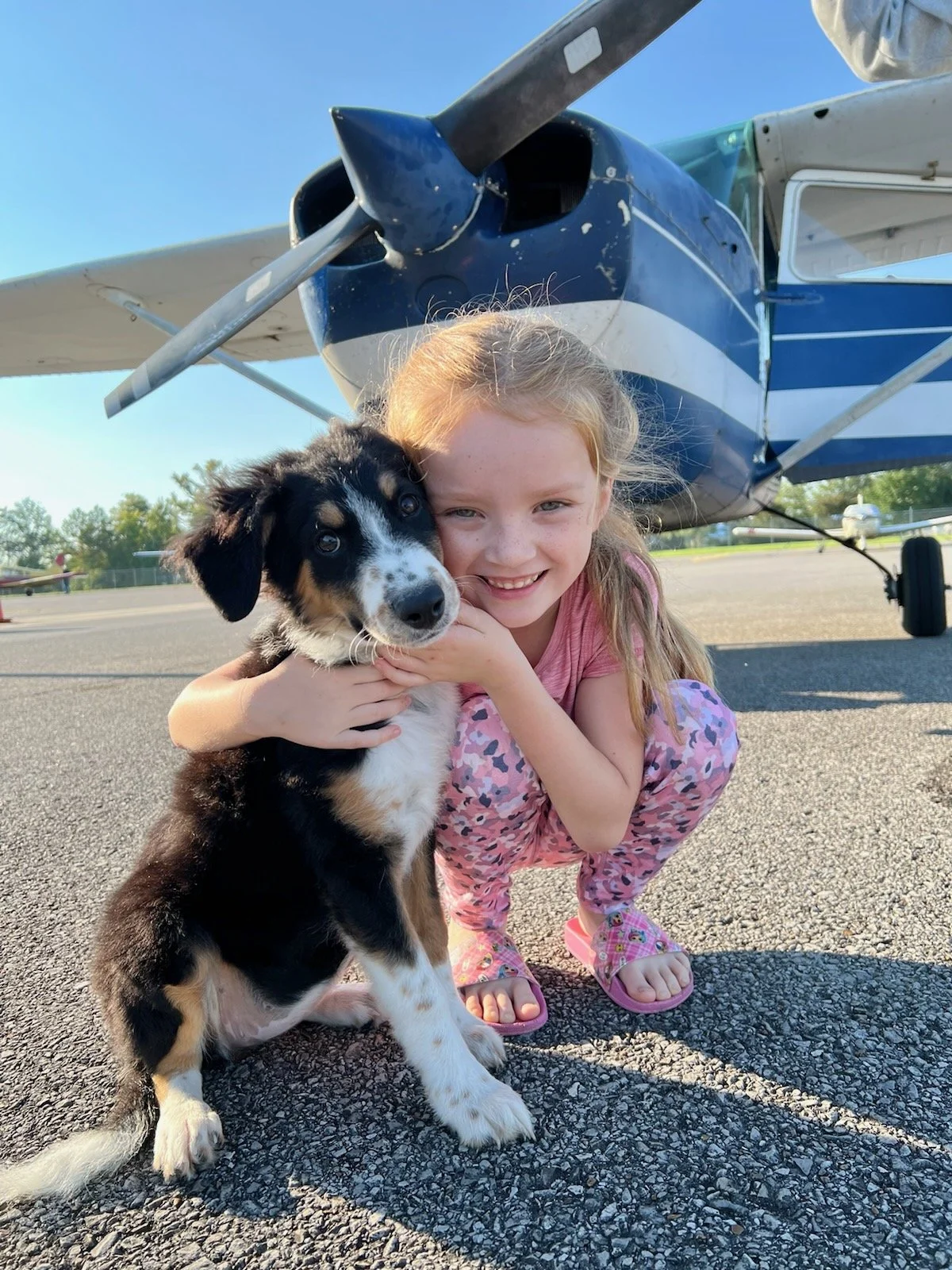 A young girl and her puppy sit on the tarmac near a small airplane, smiling and hugging during daylight.