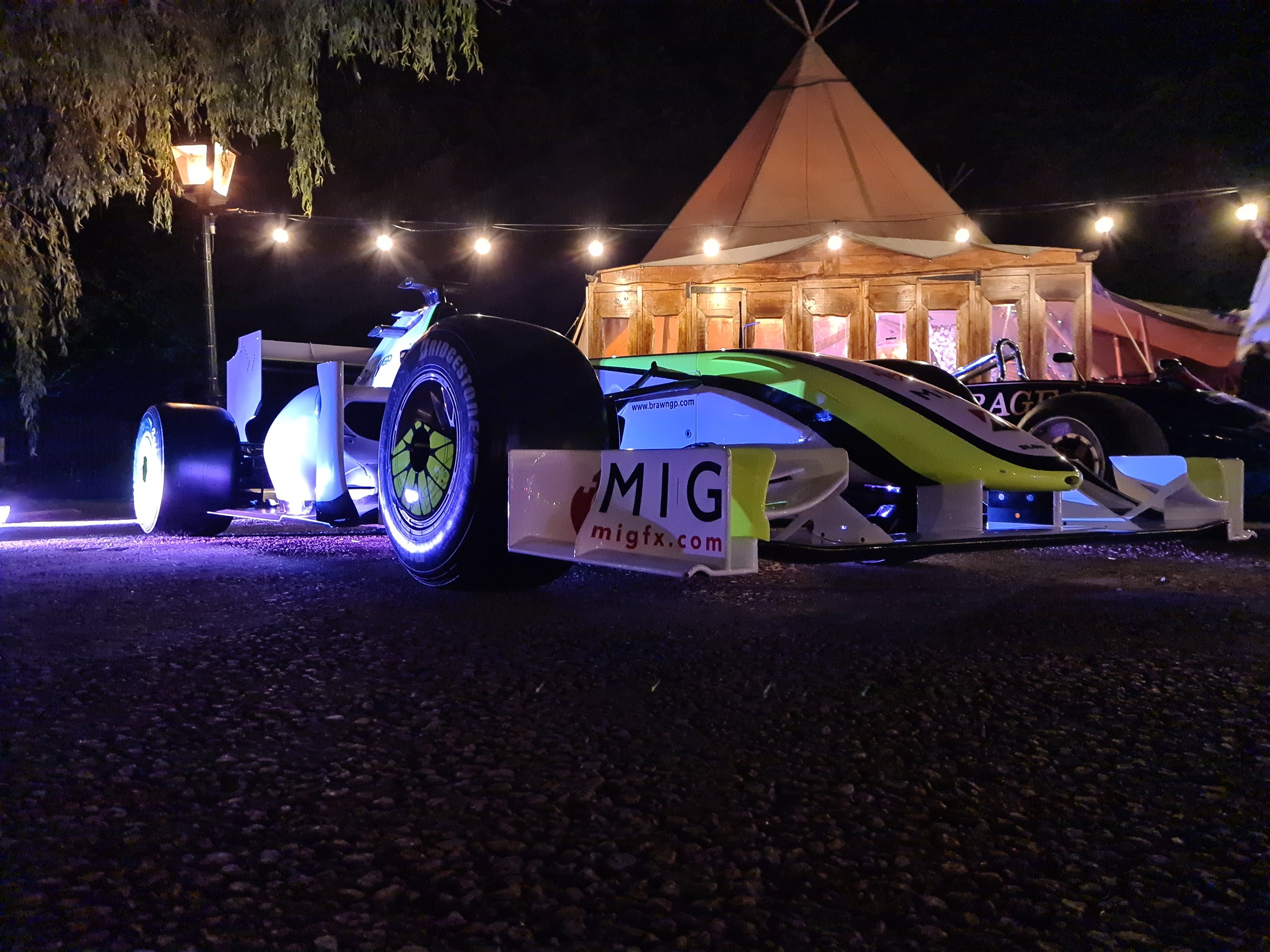 Night scene with a brightly lit Formula One car in front of a tent, surrounded by string lights and a lamppost.
