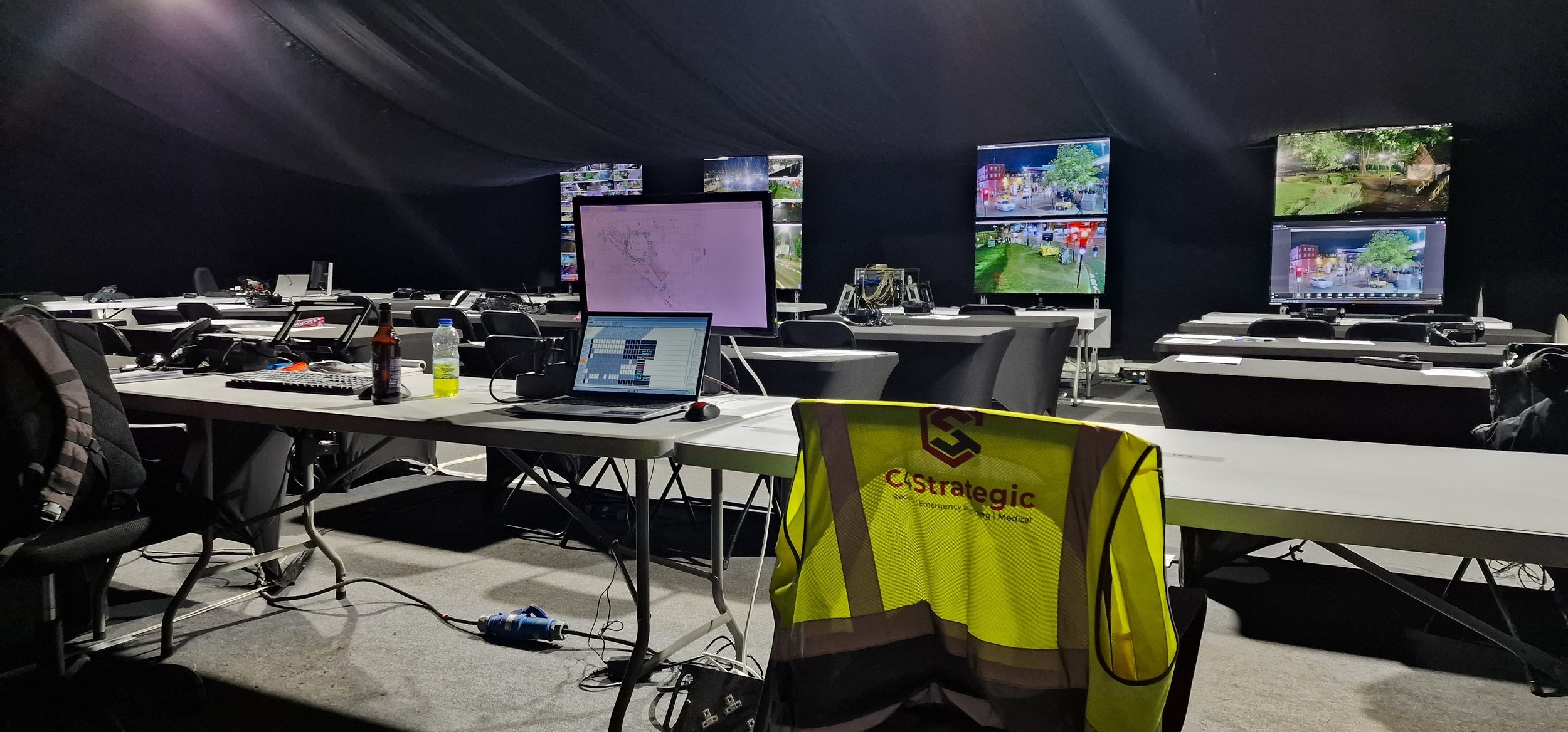 A room with rows of desks, laptops, and multiple surveillance monitors. A high-visibility jacket is draped over a chair. The setting appears to be a command or control center with open laptops, a bottle, and a can on a desk.