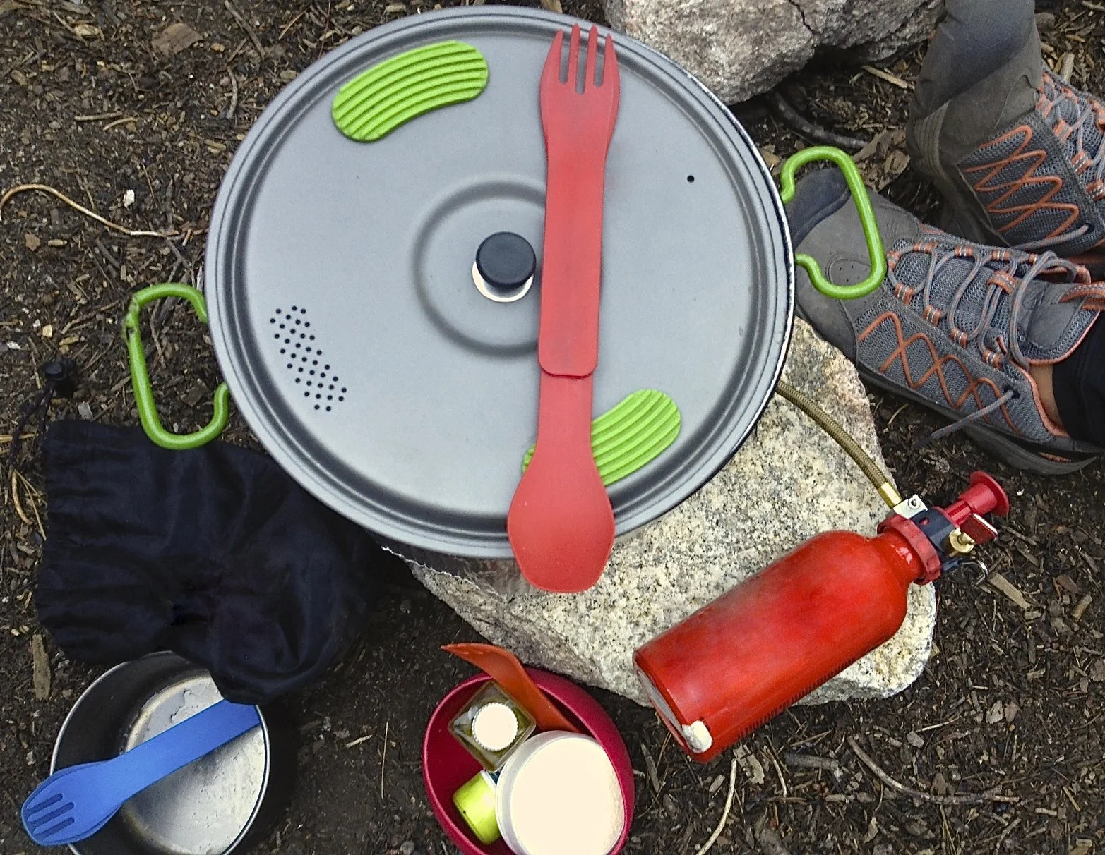 Camping gear including a pot with lid and handles, a red spork, fuel bottle, hiking shoes, and various camping utensils on rocky ground.