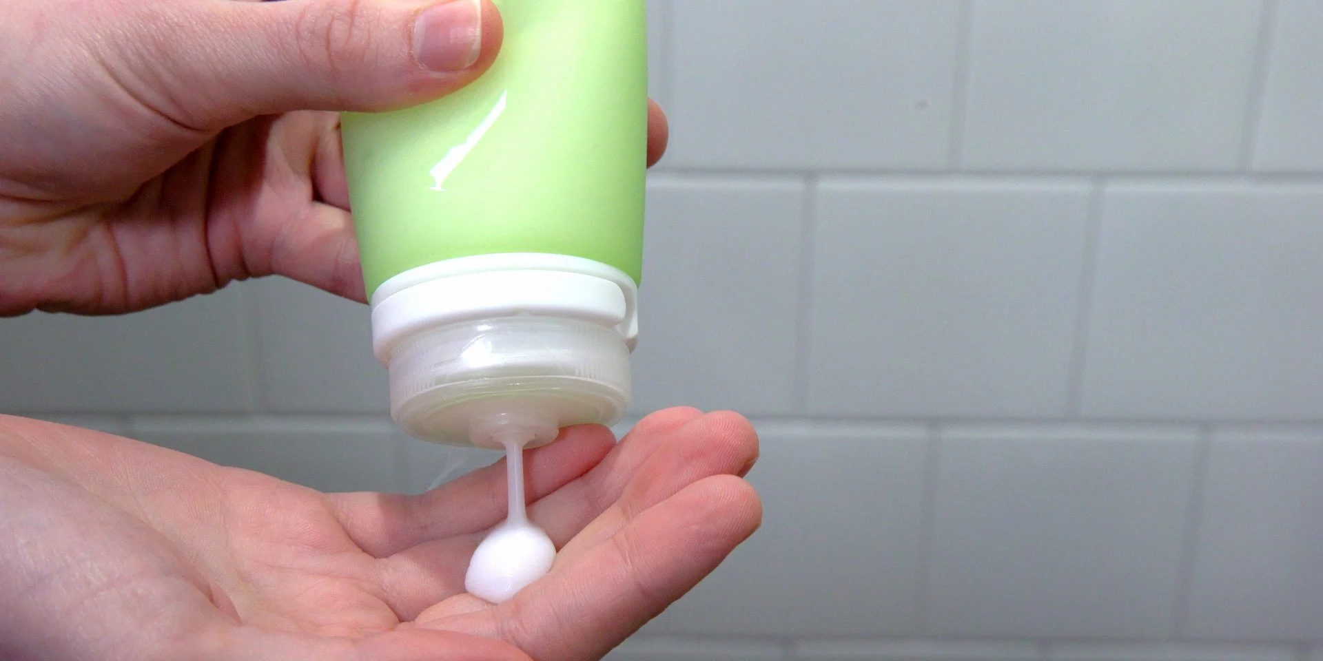 Person squeezing white lotion from a green bottle onto their palm, in front of a tiled wall.