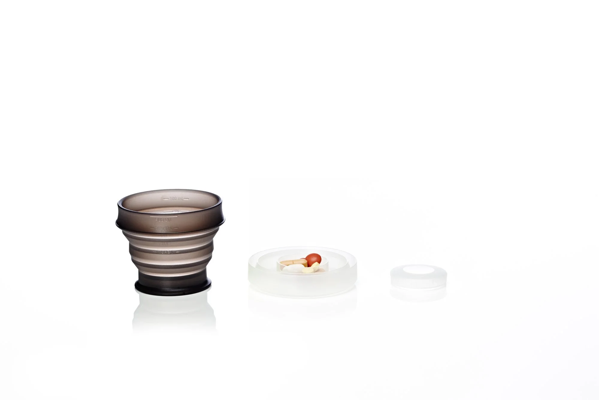 Collapsible travel cup next to a small container of mixed pills, displayed on a white surface.