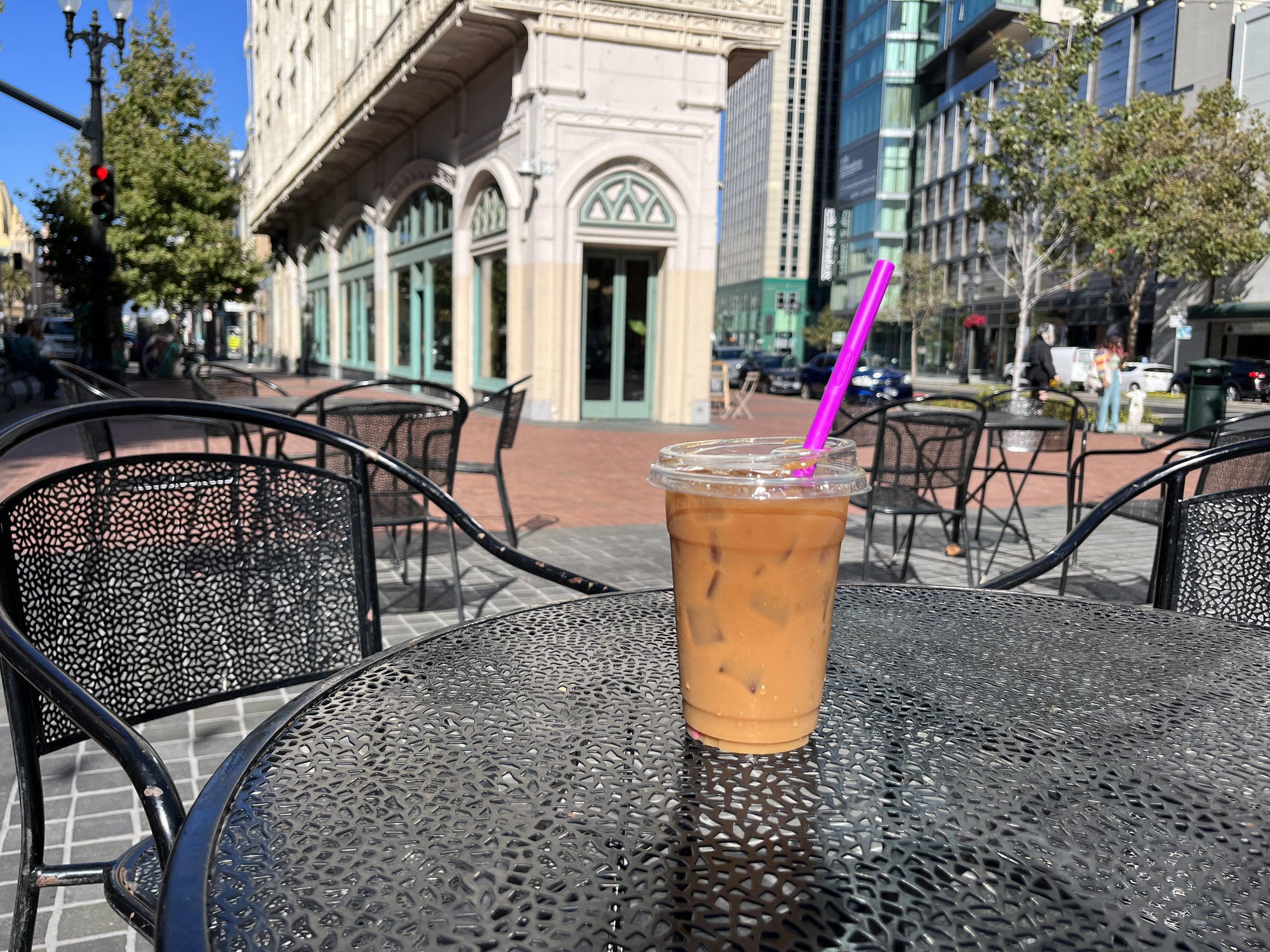 Iced coffee with a purple straw on a metal outdoor café table, with a city street and historic buildings in the background.