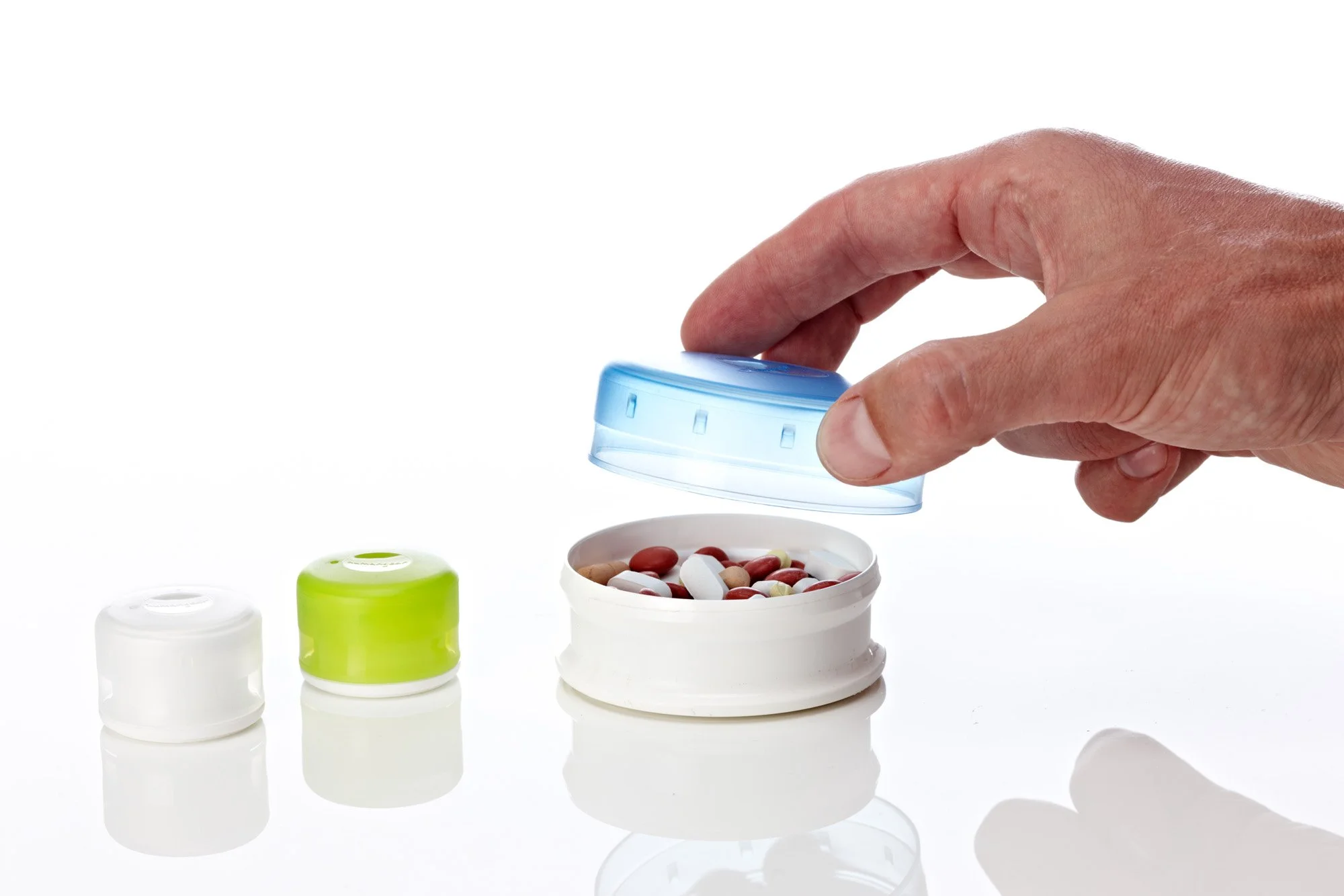 Hand lifting a blue cap from a container filled with pills, alongside two other containers with green and white caps on a white surface.