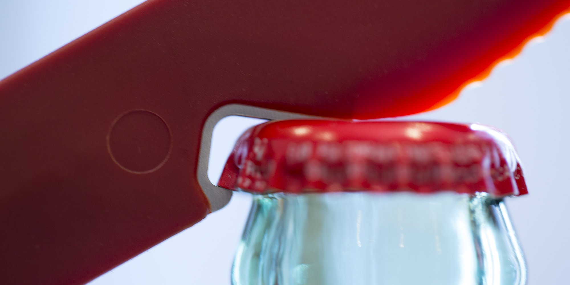Close-up of a bottle opener removing a red cap from a glass bottle.