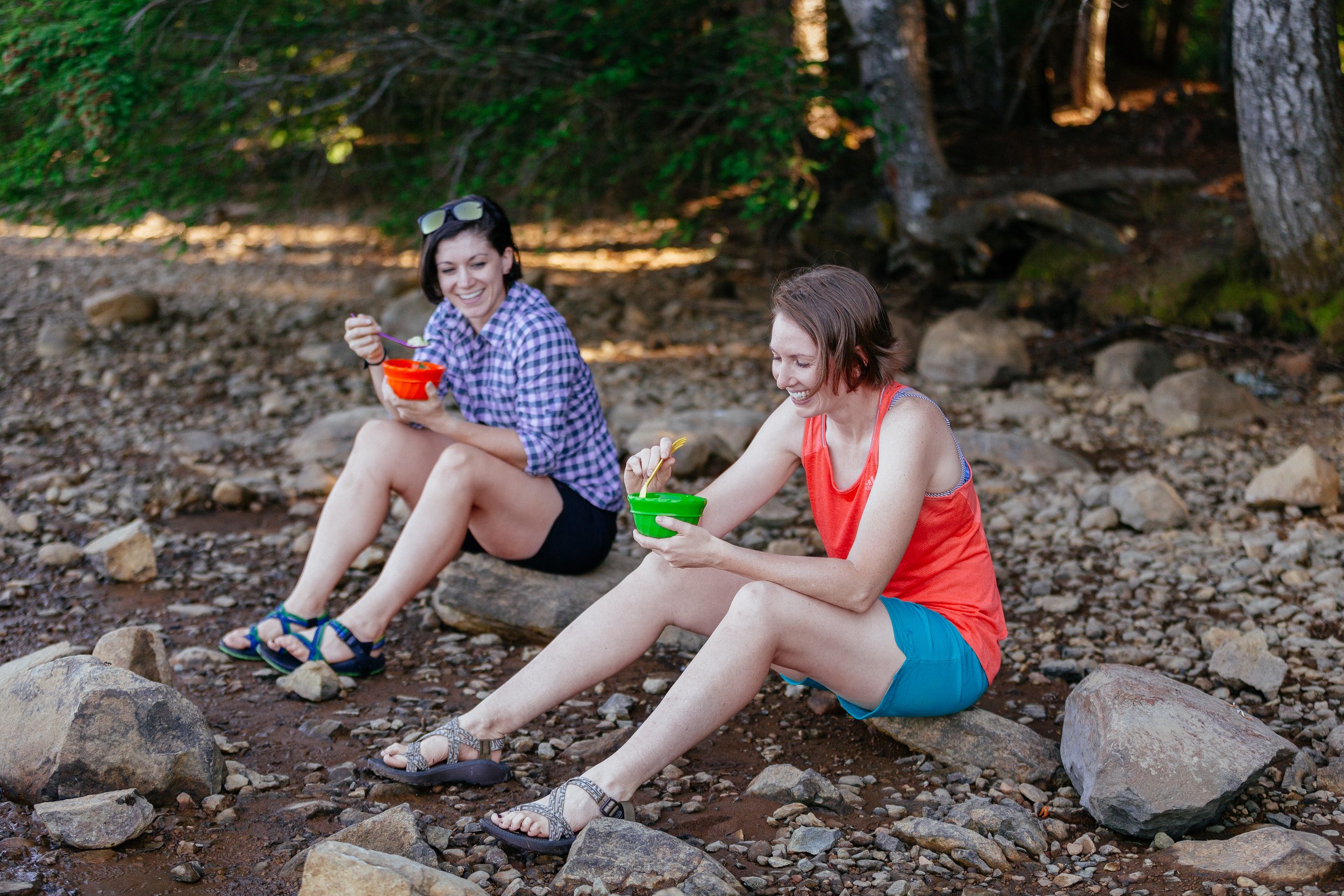 Two people sitting on rocks outdoors, eating from bowls, with a forest in the background.