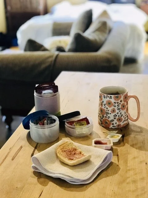Table with breakfast items including a floral mug, toast on a napkin, condiment packets, a container with tea packets, and a couch in the background.