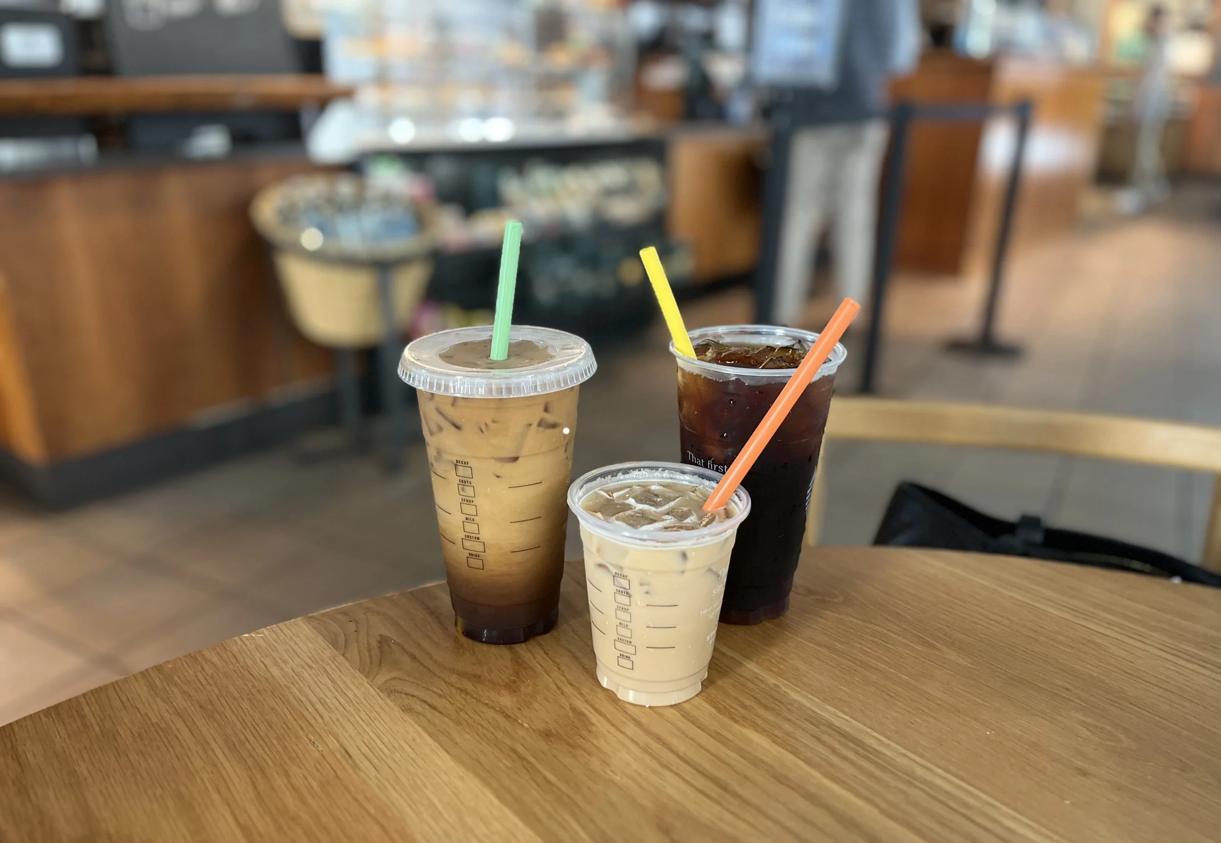 Three iced coffee beverages with colorful straws on a wooden table in a cafe.