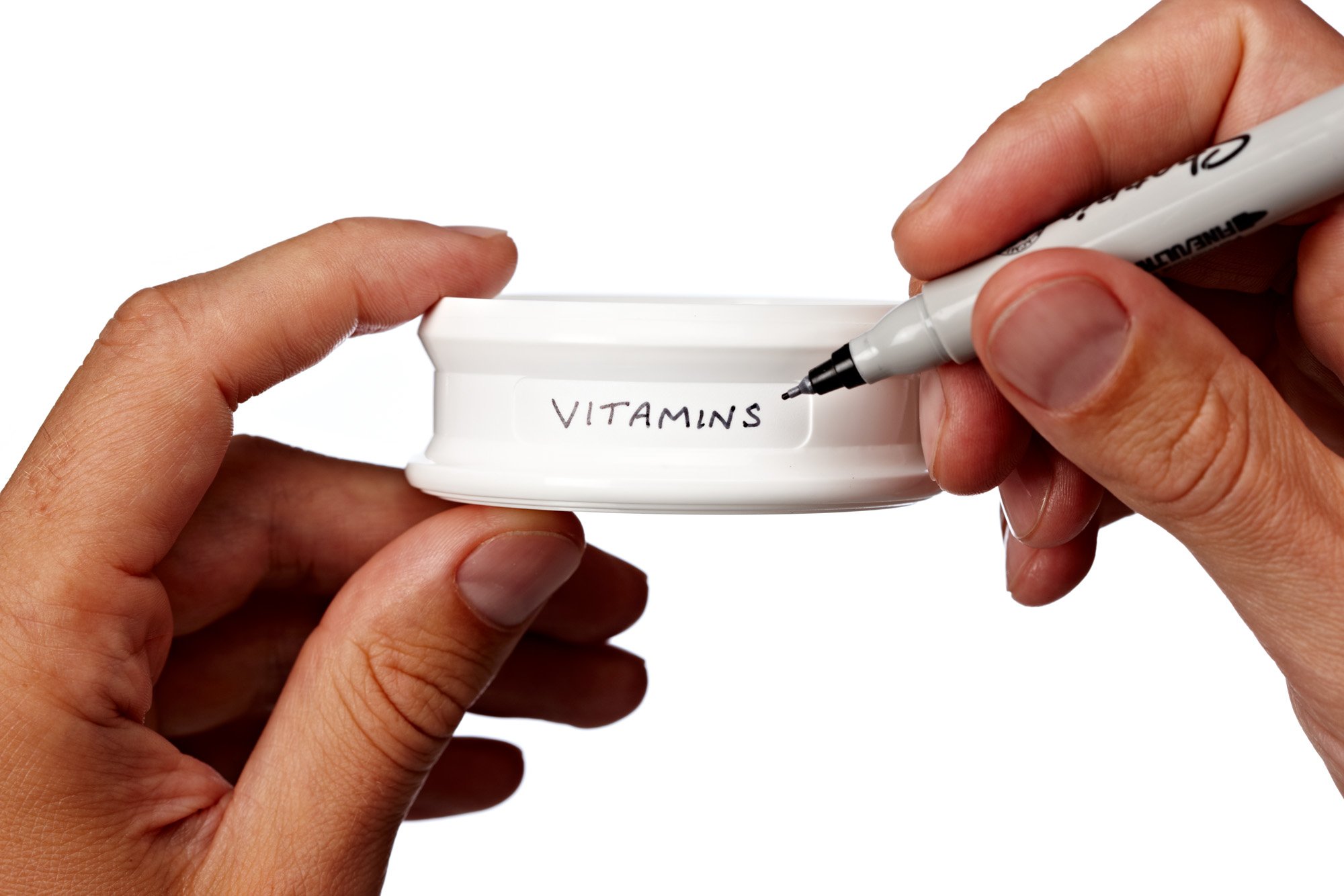 Person writing "VITAMINS" on a white container lid with a black marker