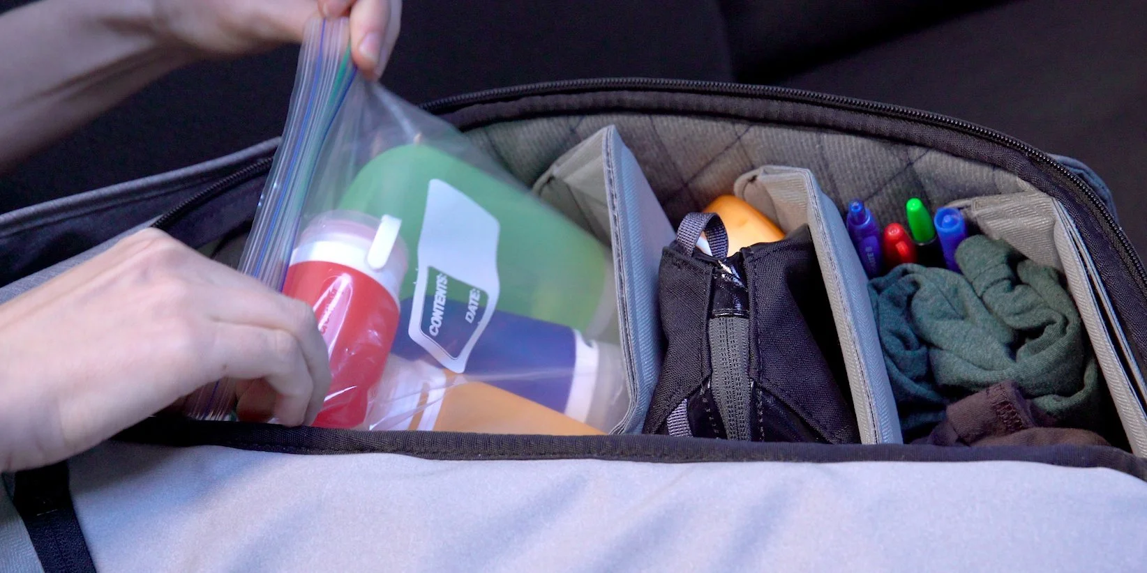 Person packing a suitcase; visible items include a plastic ziplock bag with toiletries, a small pouch, clothing, and several colored pens.