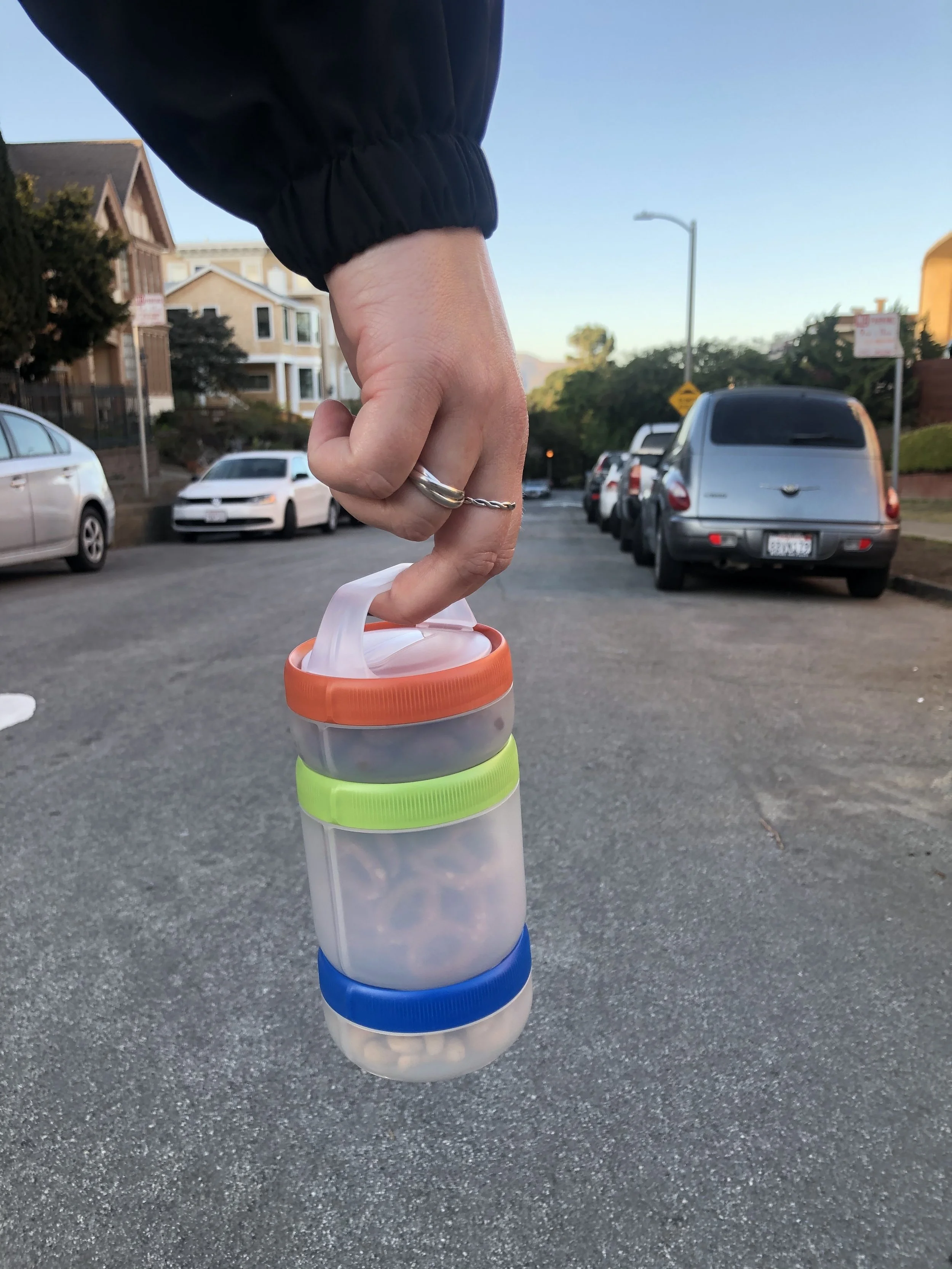 Hand holding a stackable snack container with orange, green, and blue lids, containing pretzels and nuts, on a residential street.