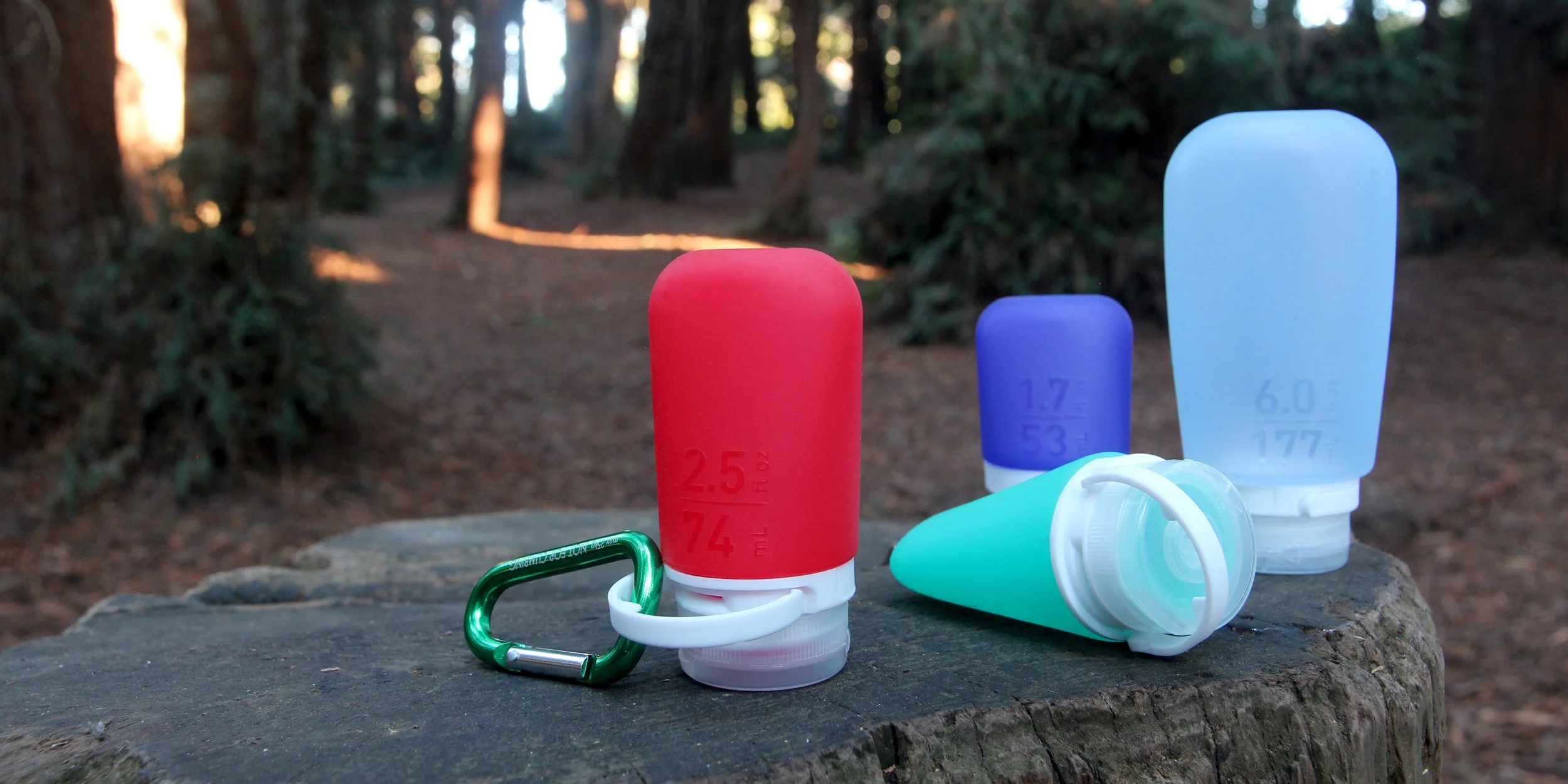Assorted silicone travel bottles with size labels on a tree stump outdoors, with a green carabiner.