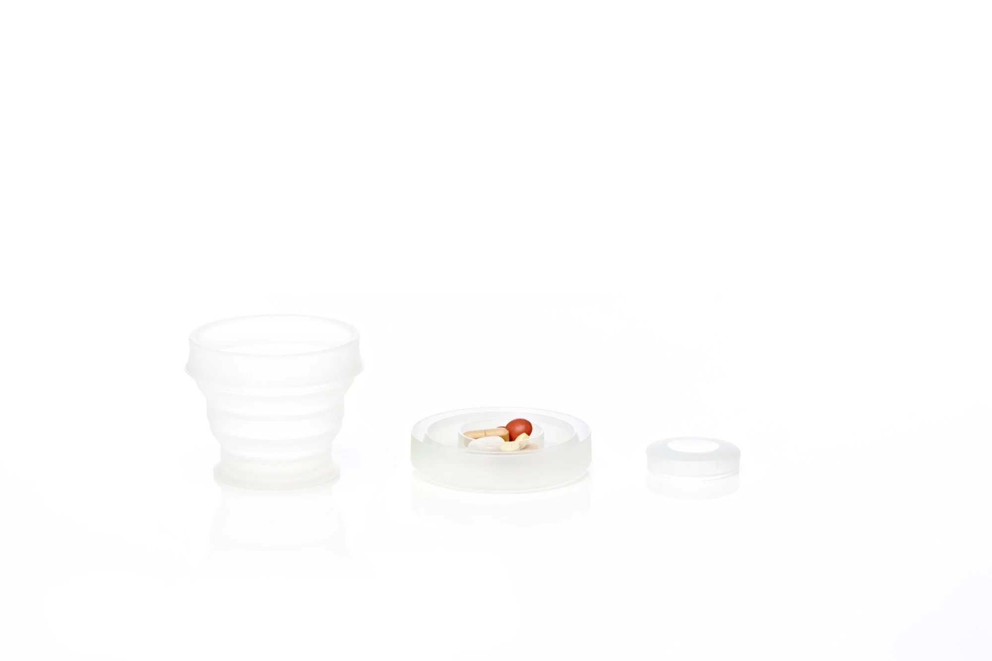 Collapsible silicone cup, pill container with assorted pills, and a silicone lid on a white background.