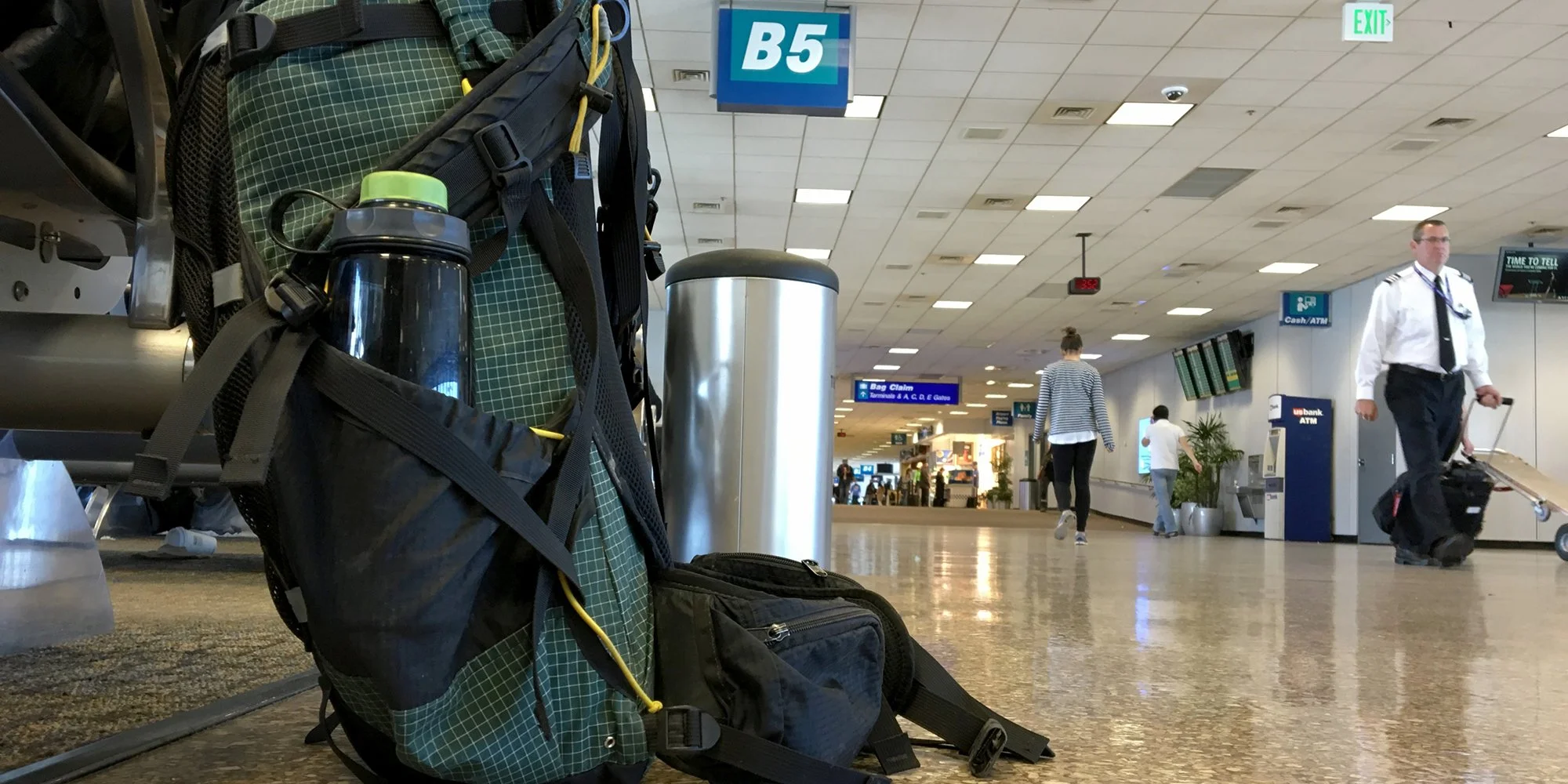 Backpack and water bottle on airport floor near gate B5, with passengers and crew walking by.