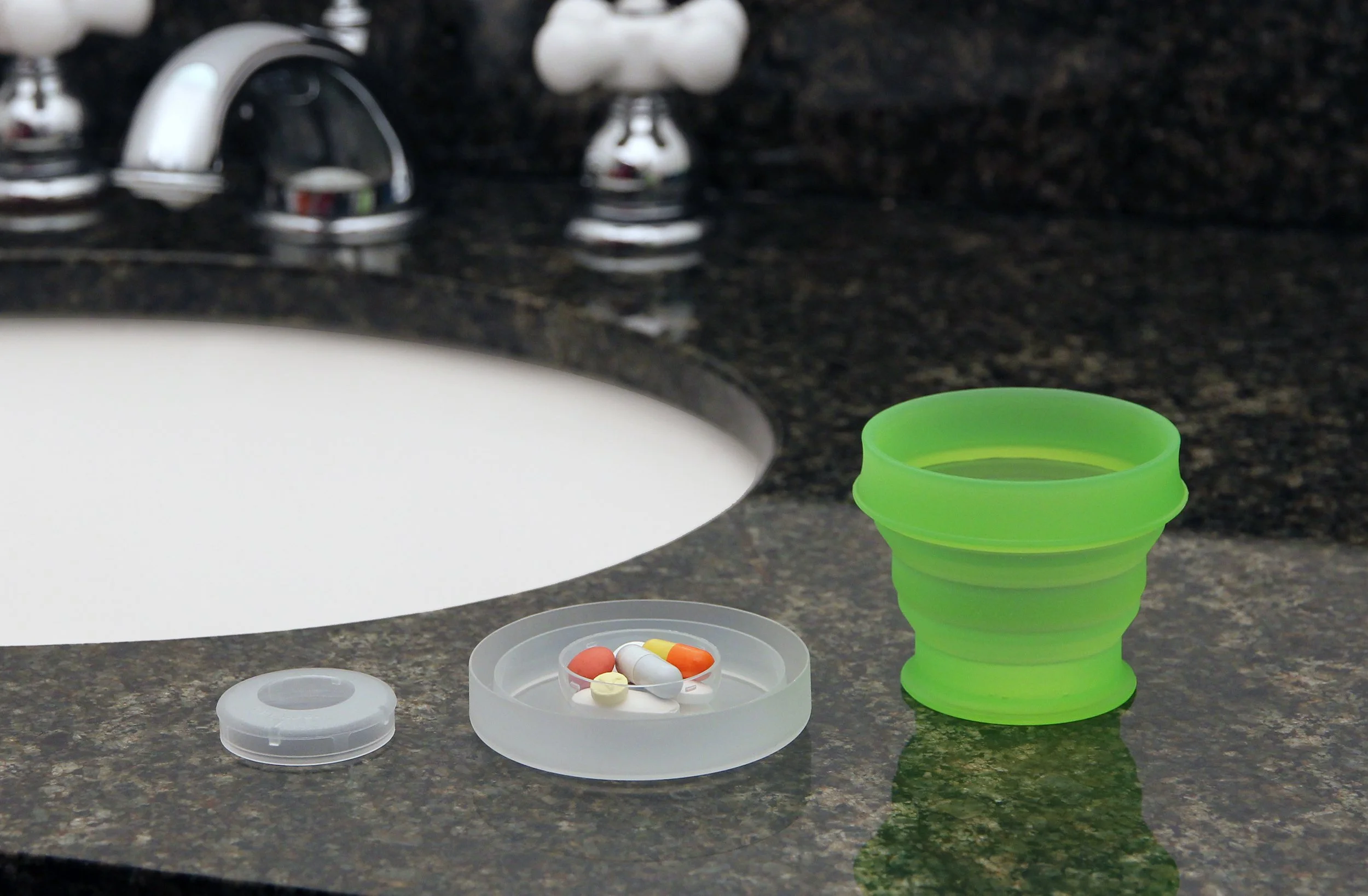 Close-up of a bathroom sink with a green collapsible cup filled with water, and a round container holding various pills on the counter.