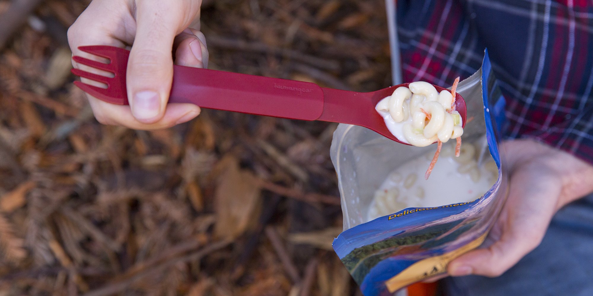 Person holding a red spork with macaroni salad over an open food pouch outdoors.